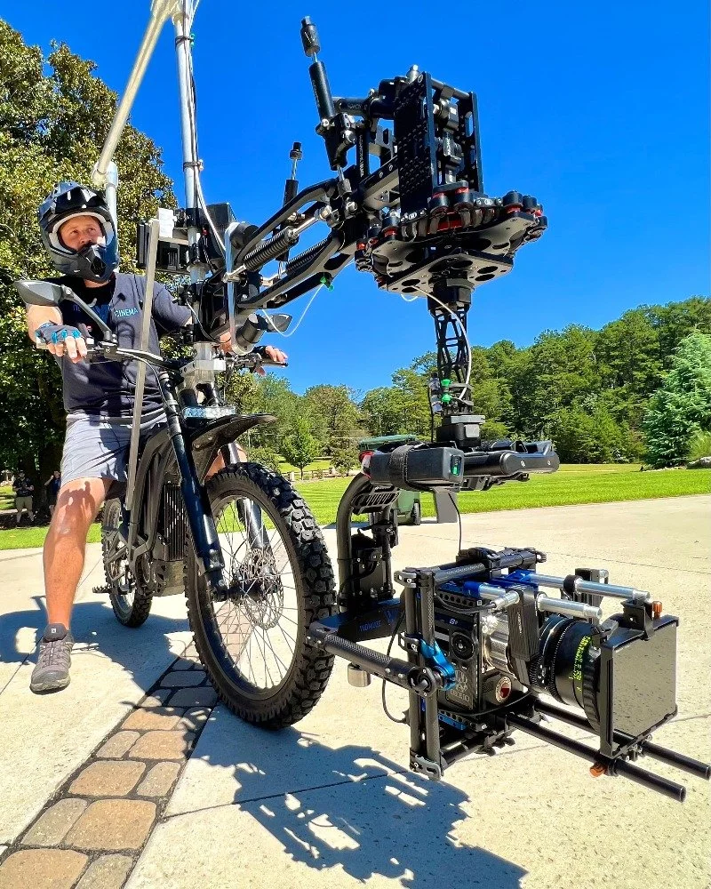 A person wearing a helmet and cycling gear sitting on a mountain bike, equipped with a large, complex camera rig mounted on the front, outdoors on a sunny day with trees and a park in the background.