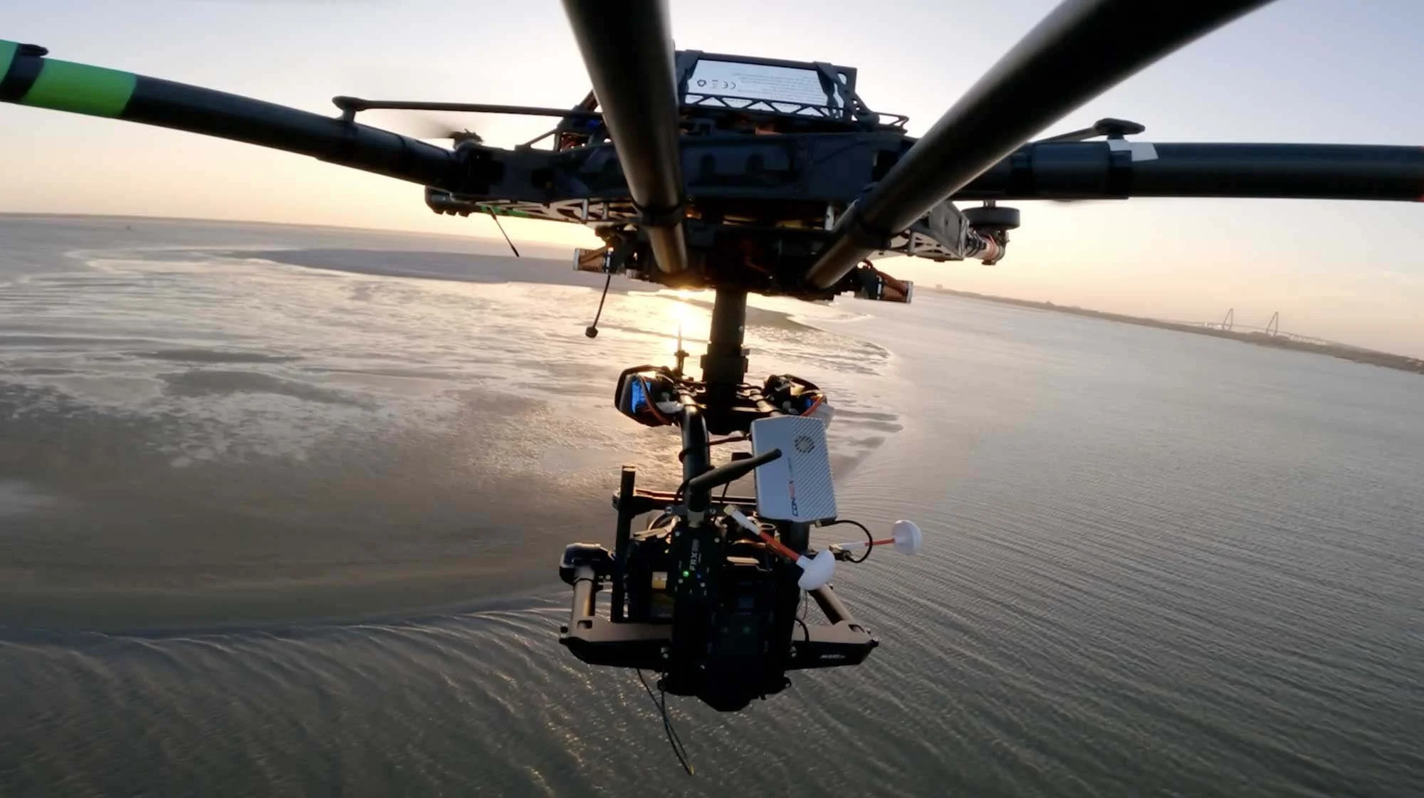 A drone flying over a body of water during sunset, with a bridge visible in the distance.
