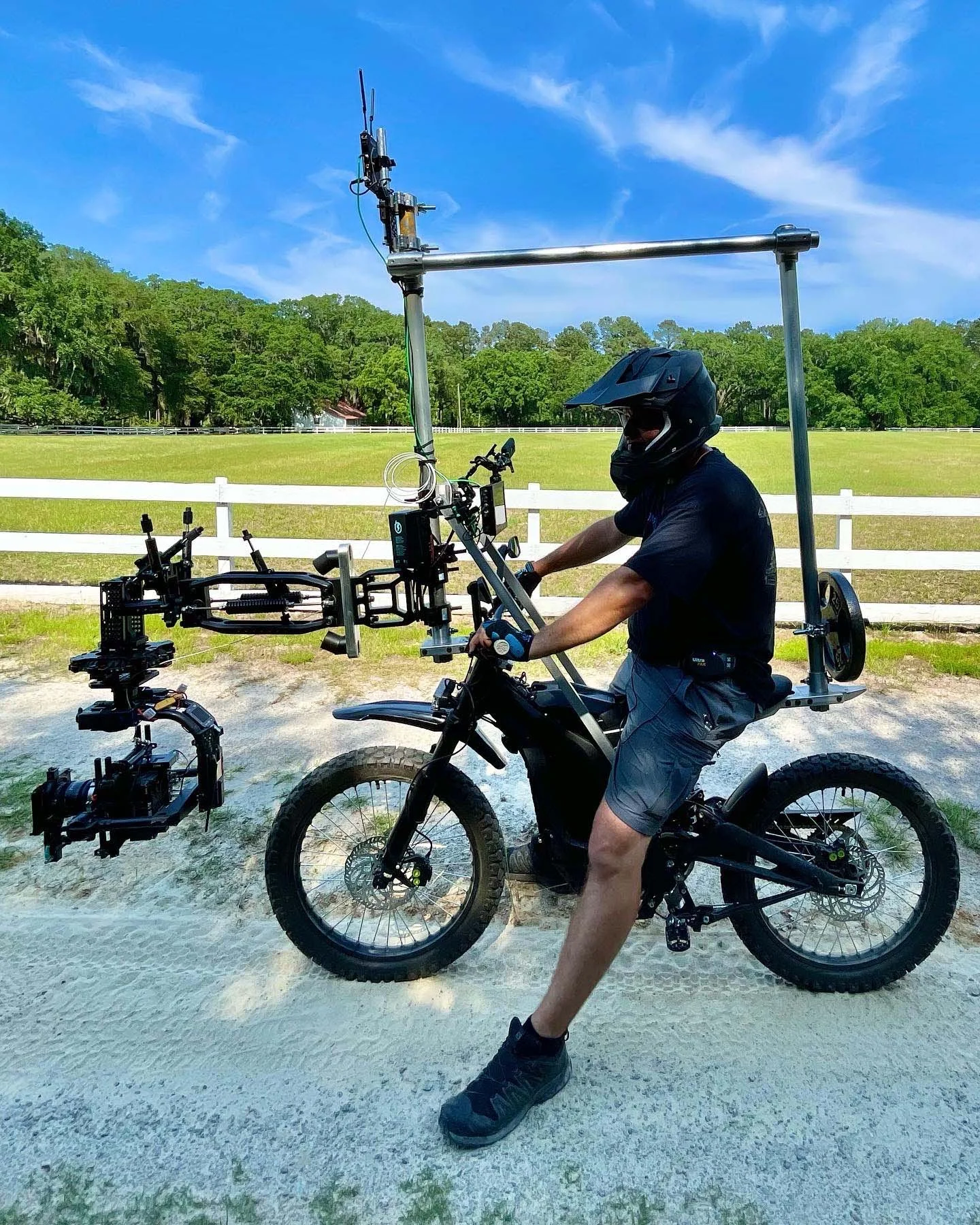 Person riding a mountain bike equipped with a camera stabilizer rig and mounted camera, wearing a helmet and gloves, in a rural outdoor setting with a white fence and green trees in the background.