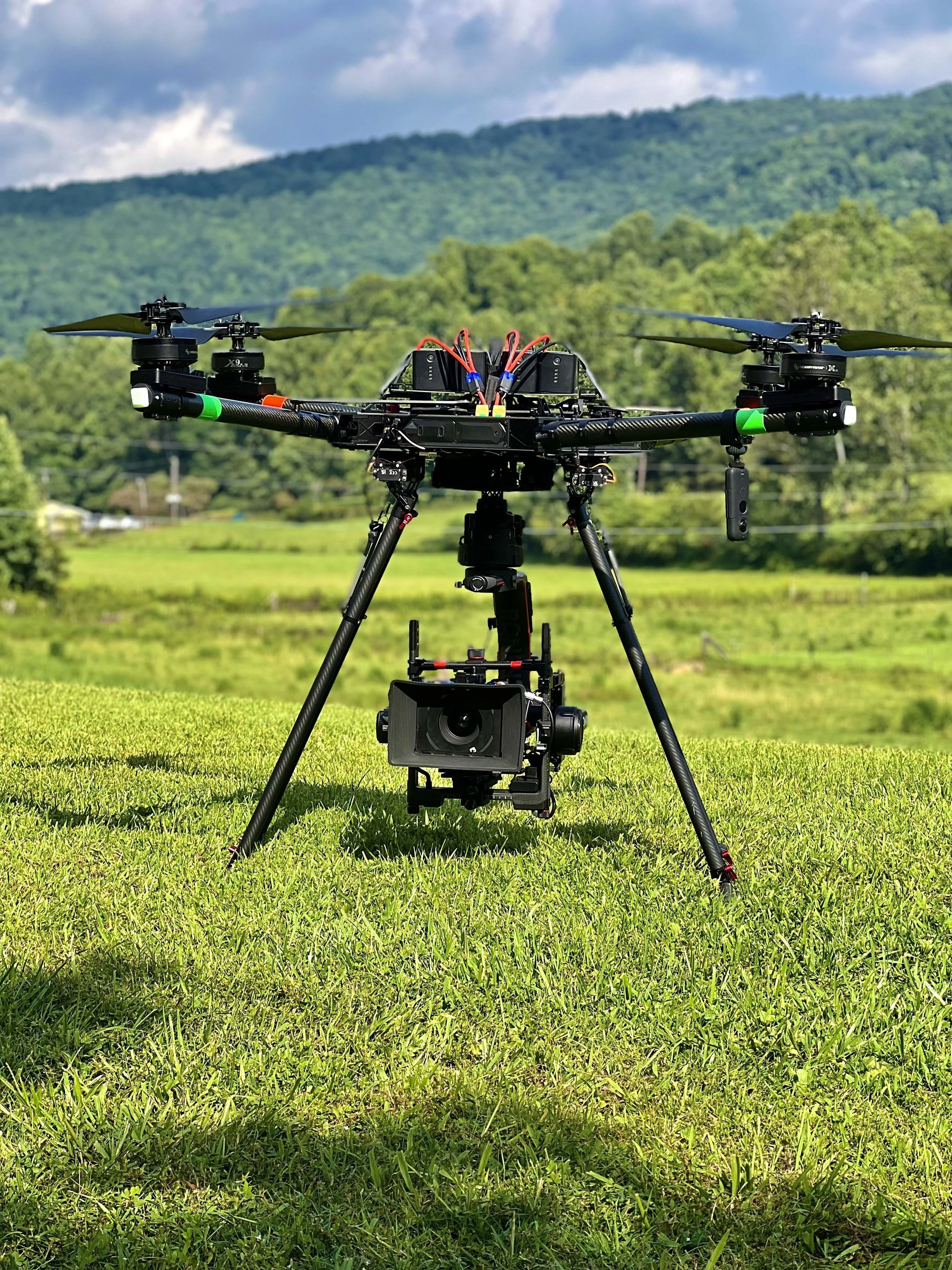 A drone equipped with a camera is positioned on grass with a green landscape and mountains in the background.