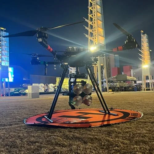 A military drone with four propellers and multiple missile payloads attached to its underside, situated on a red and black landing pad outdoors at night with illuminated structures in the background.