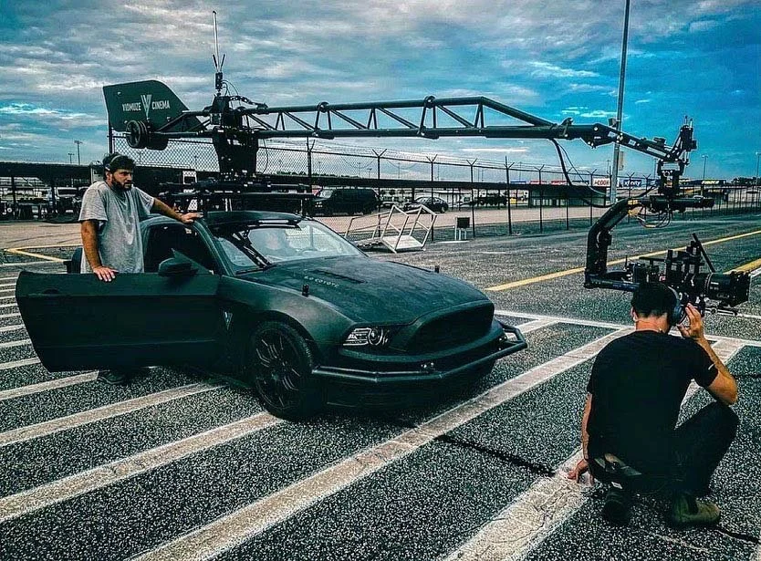 Behind the scenes of a film or commercial shoot, a man stands next to a matte black car with a camera operator filming from a low angle, and a large camera crane is positioned over the car in an empty parking lot.