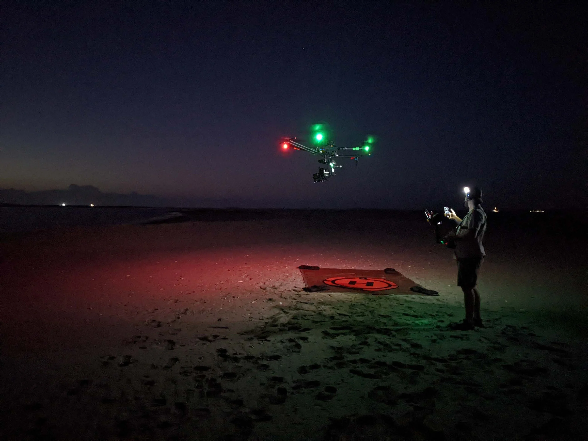 A person operating a remote-controlled drone at night on a sandy beach with an illuminated landing pad; the drone has green and red lights and is flying above the beach.