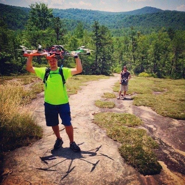 A man in a neon green t-shirt and navy shorts holding a drone above his head outdoors on a rocky trail. A woman with a backpack stands in the background, surrounded by trees and mountains.