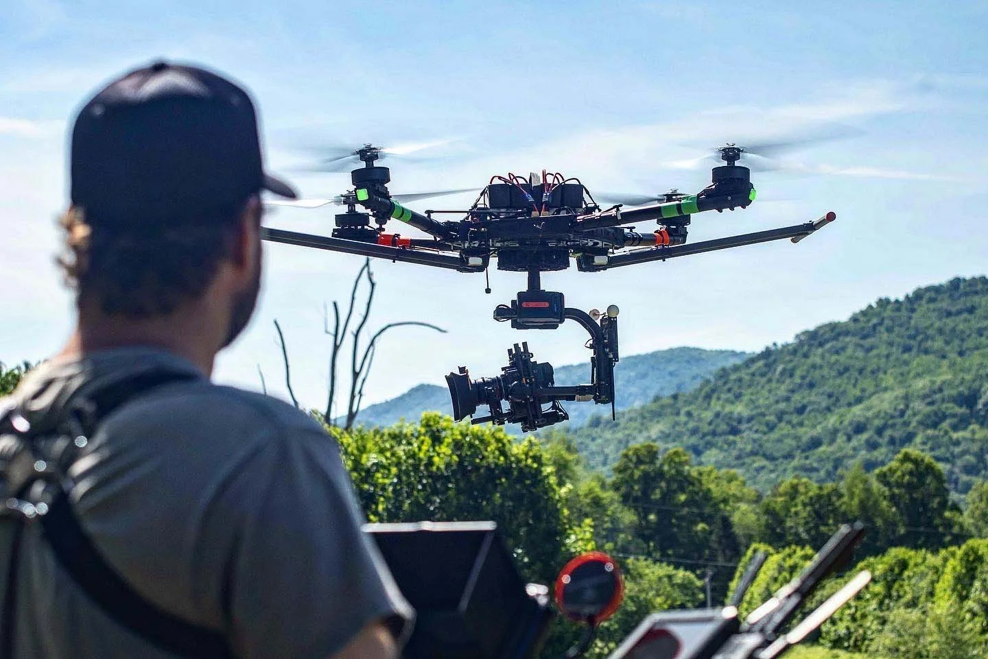 A drone with a camera mounted on it flying outdoors with a person in the foreground operating equipment, against a backdrop of green hills and a blue sky.