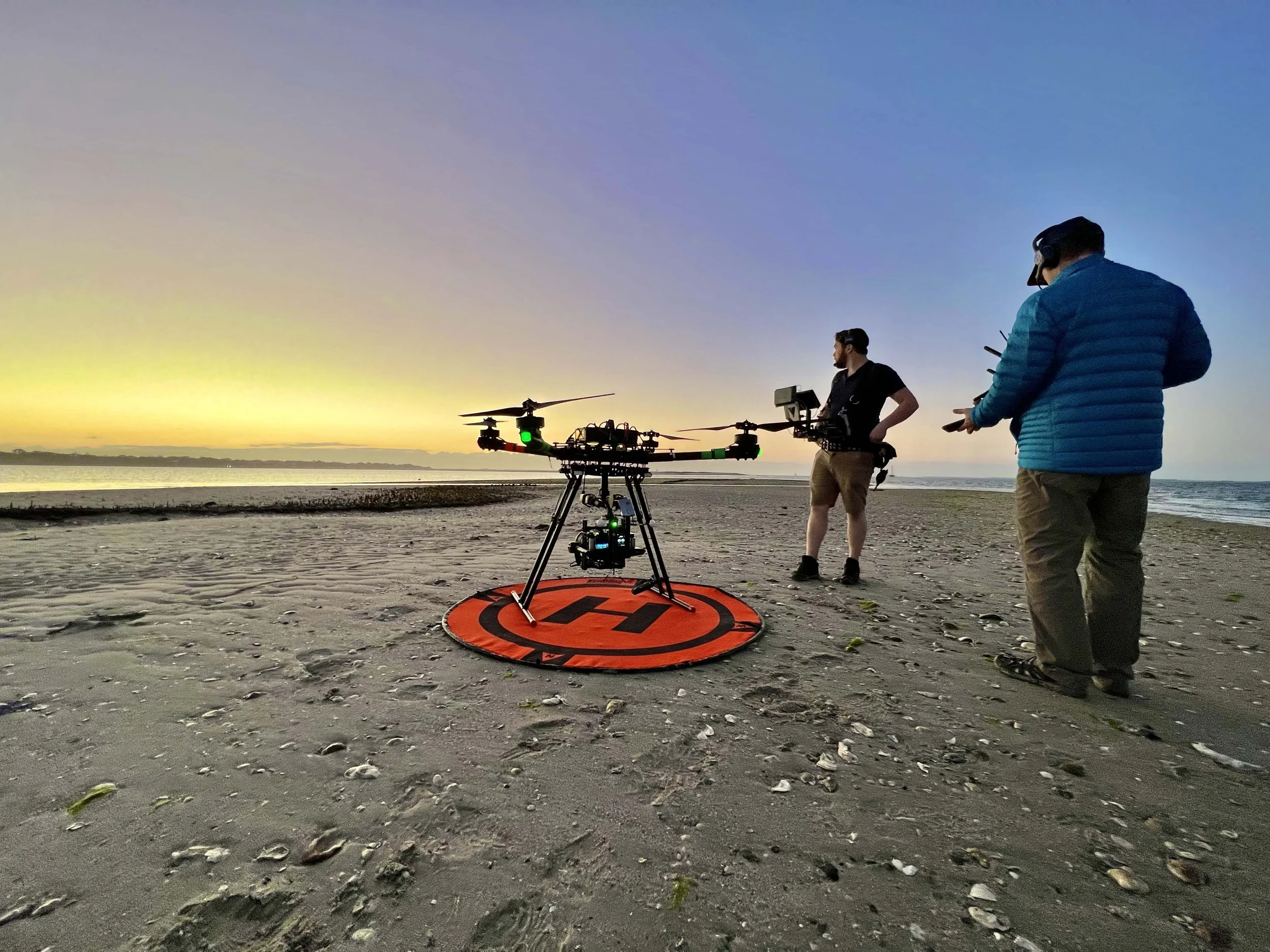 Two people operating a drone on a beach at sunset. One person is working with a monitor, and the other is holding a remote control. The drone is on a landing pad in the sand.