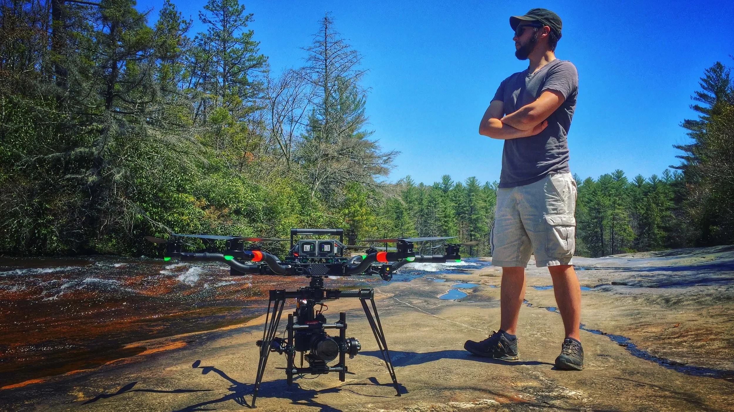 A man stands with arms crossed next to a large drone on a rock formation beside a river, with trees and a blue sky in the background.