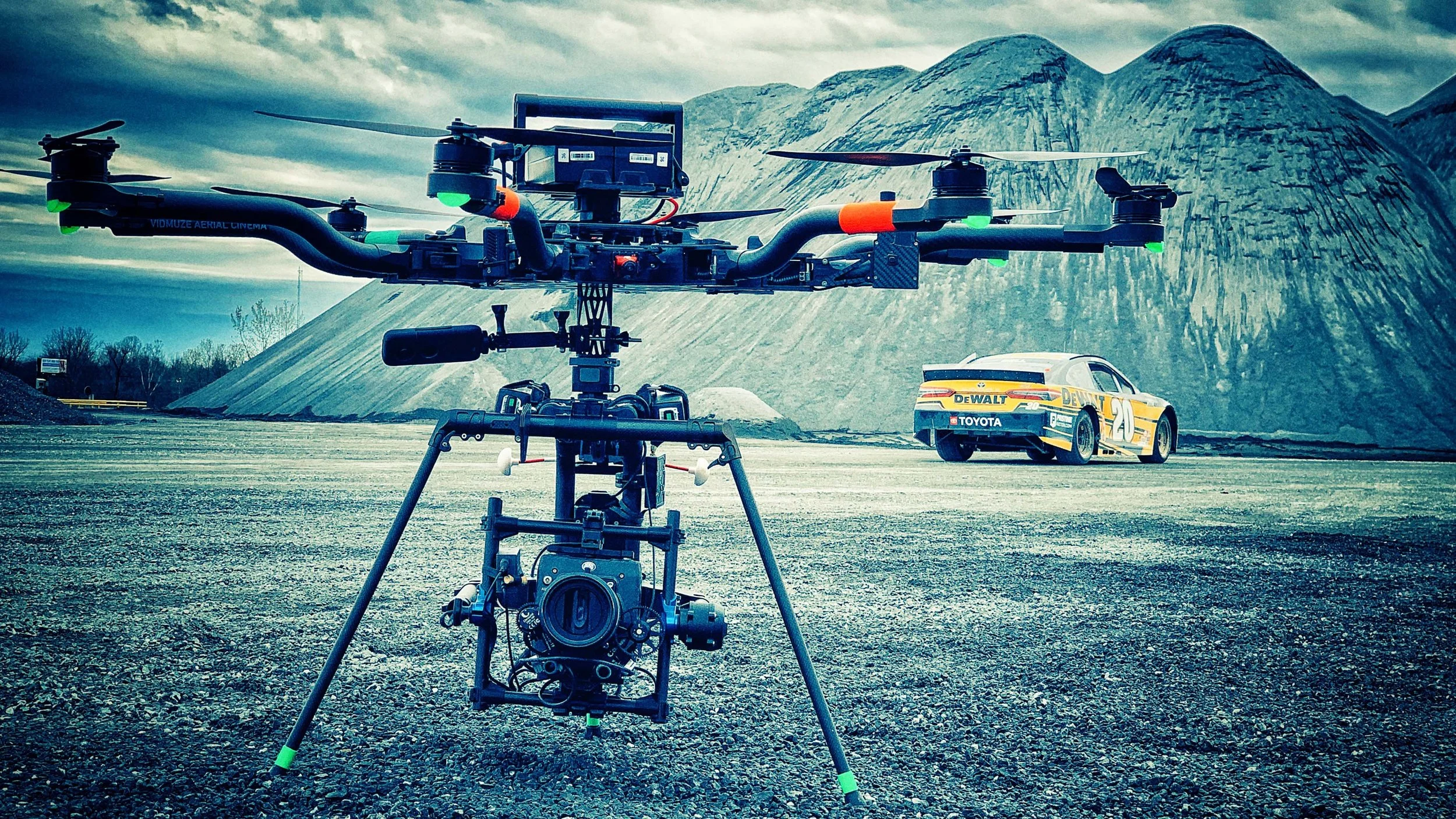 A professional camera drone with a camera mounted underneath, situated on a gravel surface outdoors, with a racing car and mountainous landscape in the background.
