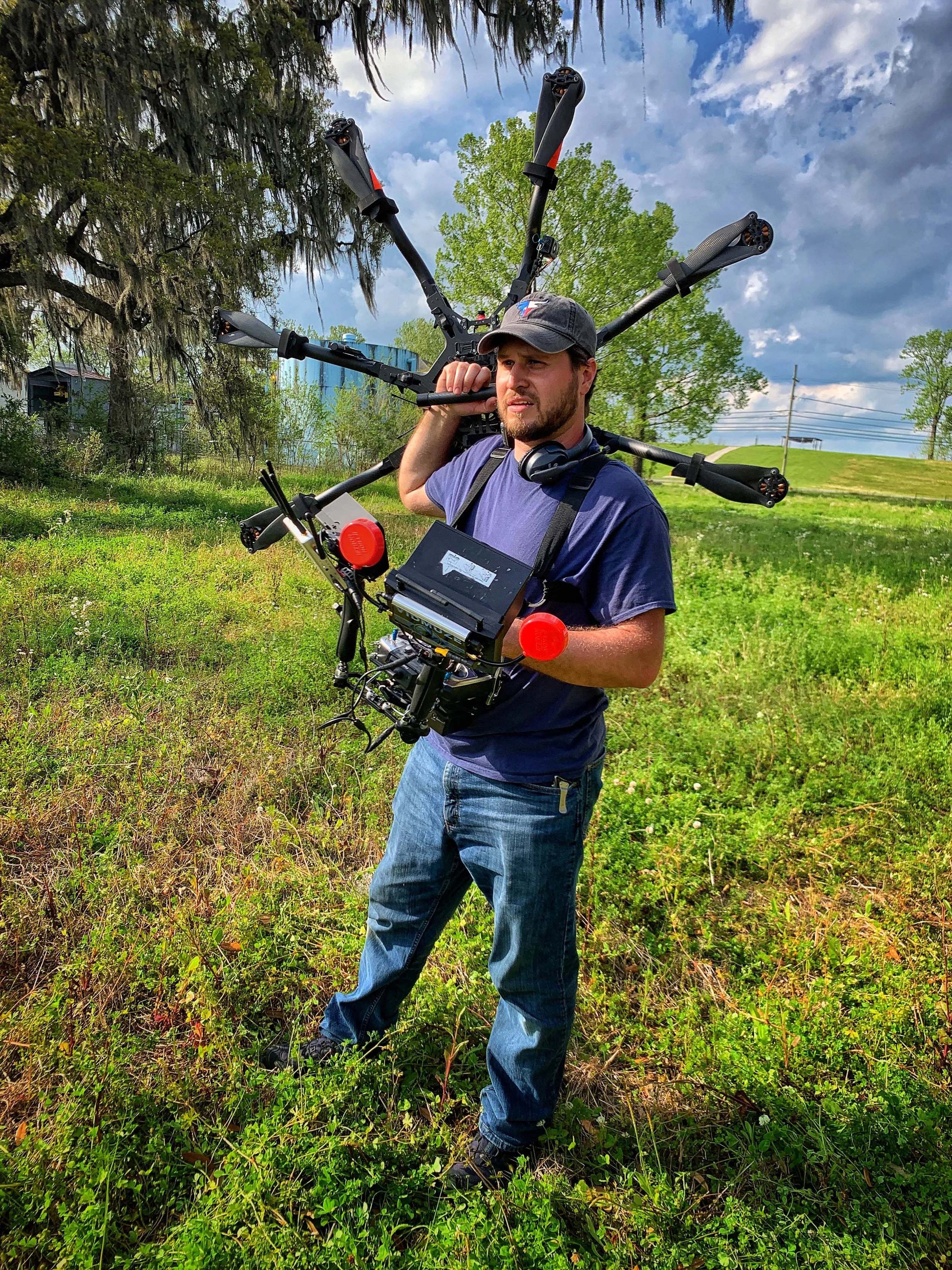 A man standing in a grassy outdoor area with trees and a cloudy sky, wearing a hat, headphones, and carrying a drone with multiple arms and camera equipment.