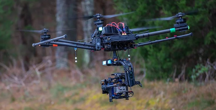 A large drone with multiple propellers flying in a natural outdoor setting with green trees and grass.