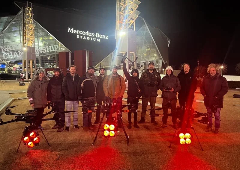 Group of people standing outside Mercedes-Benz Stadium at night, with three large drones equipped with lights in the foreground.