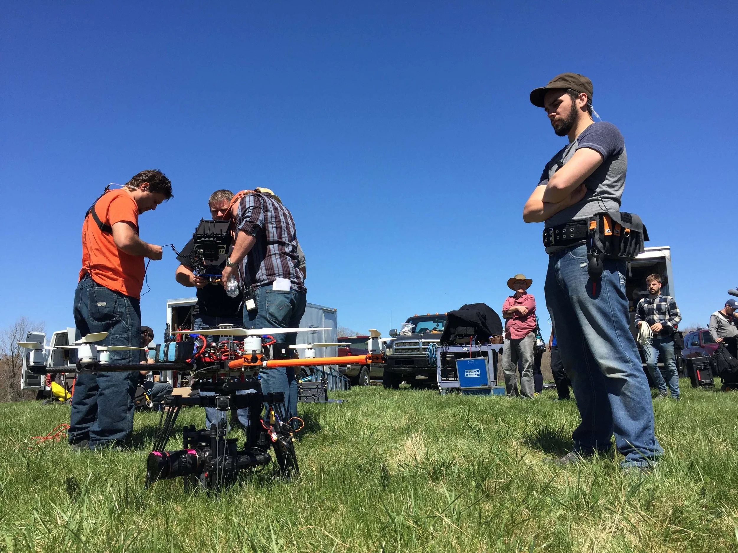 A group of people working on a large drone outdoors on a bright, clear day with a blue sky. Some individuals are focused on the drone, while others observe from the background.