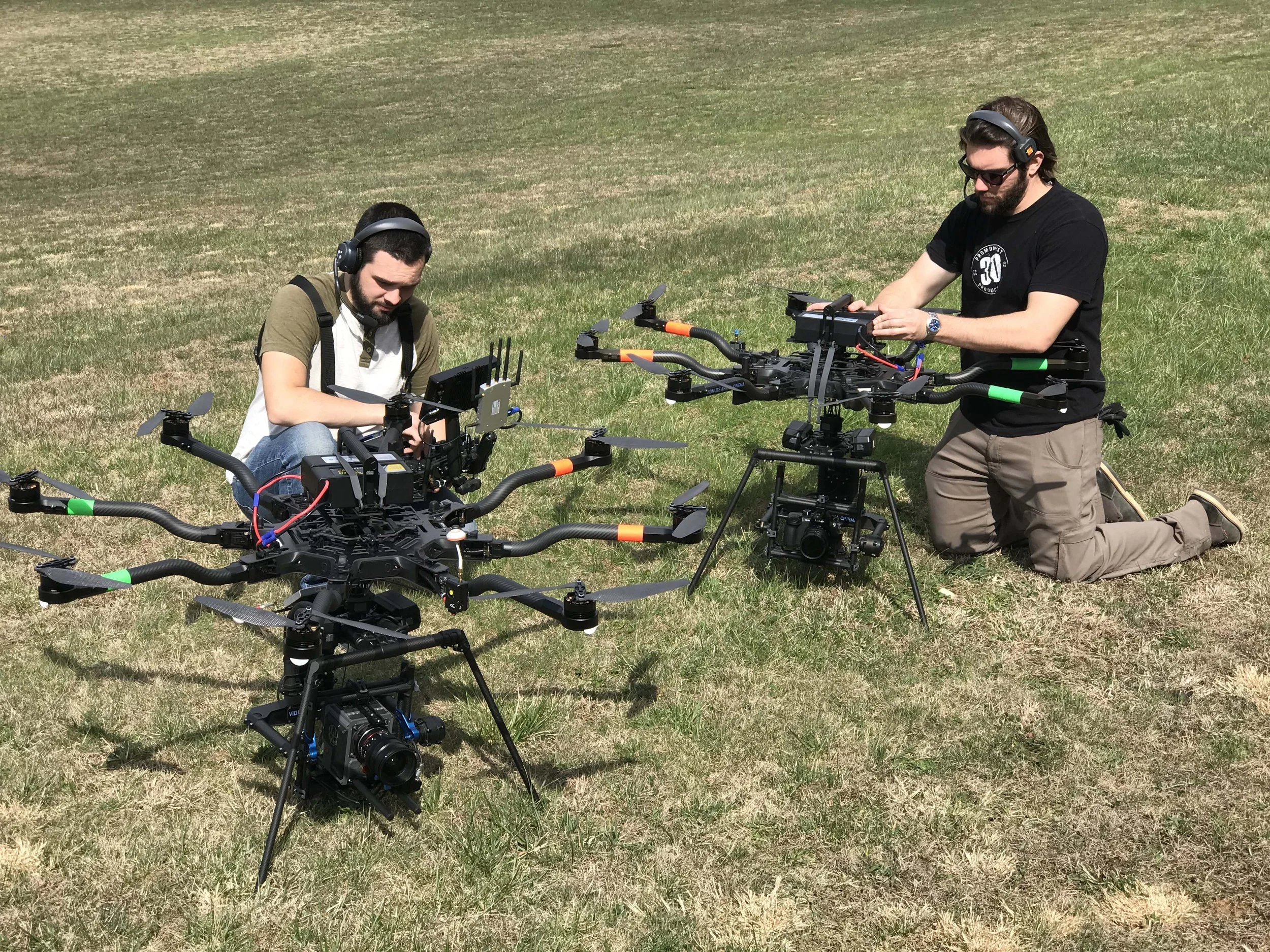 Two men working on large drones outdoors on grass, with one kneeling and the other standing, both wearing headphones and safety glasses.