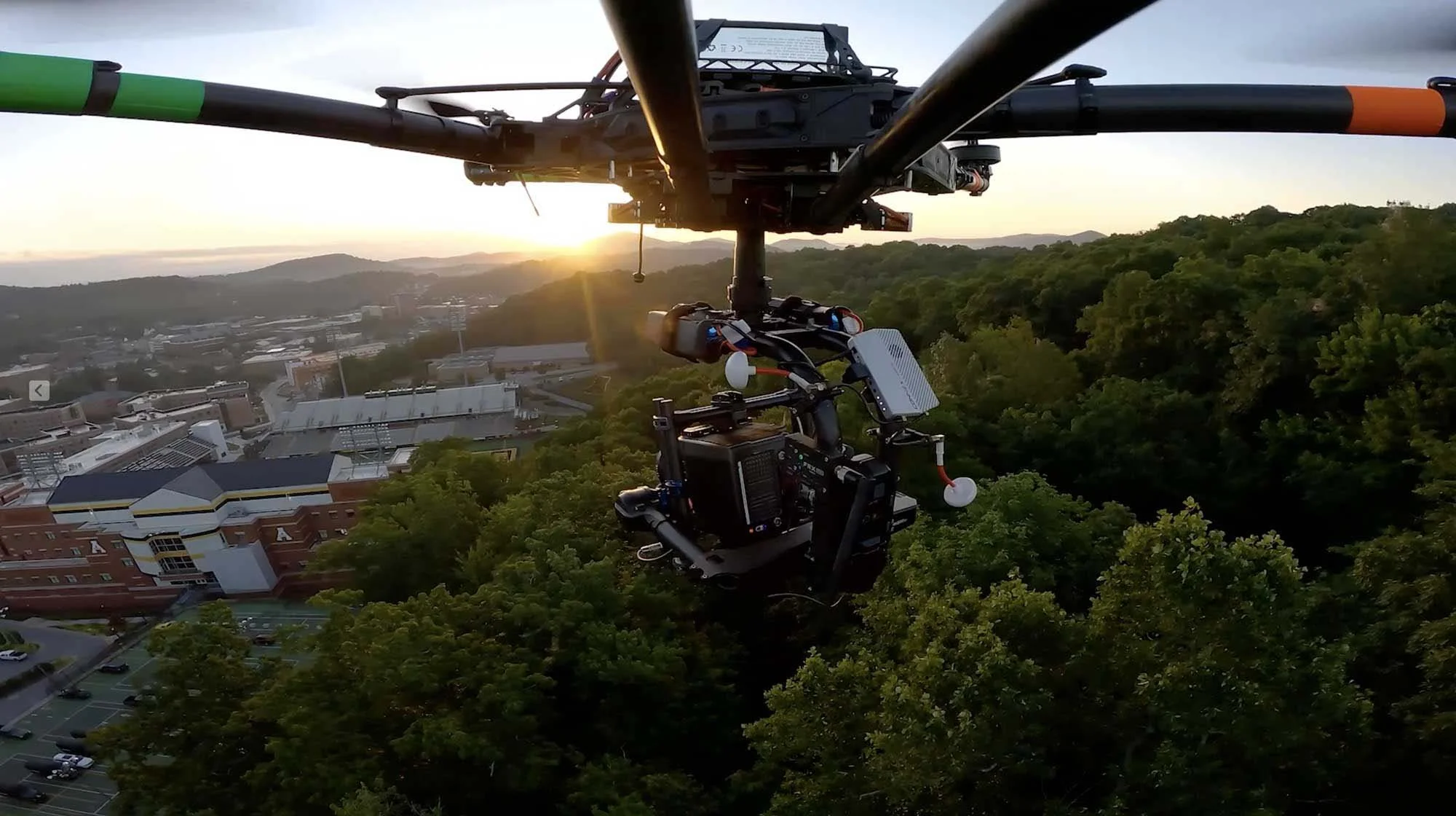 A drone flying over a cityscape during sunset, with buildings, trees, and distant hills visible.