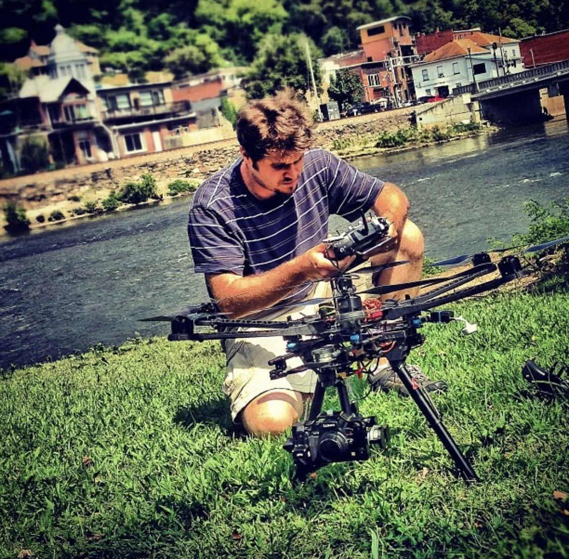 A man kneeling on grass beside a river, holding a remote control, with a drone on the ground in front of him, and a cityscape with colorful buildings and bridges in the background.