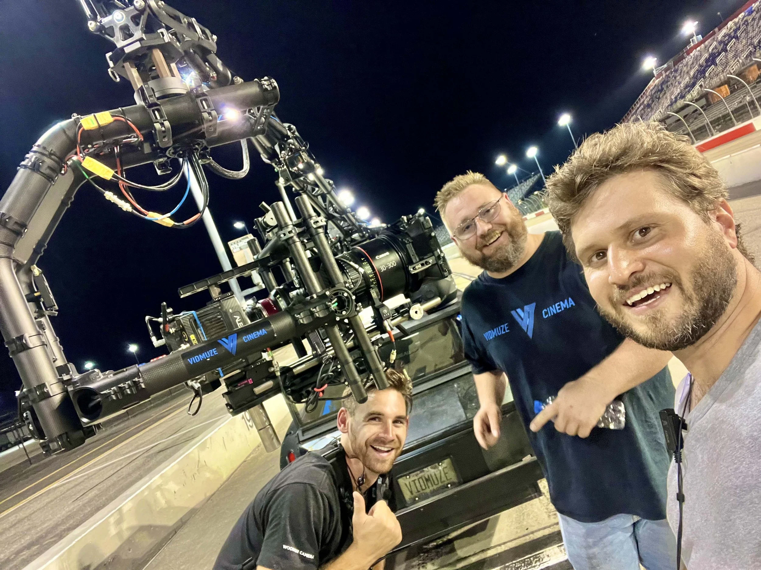 Three men taking a selfie next to a large camera rig on a race track at night.