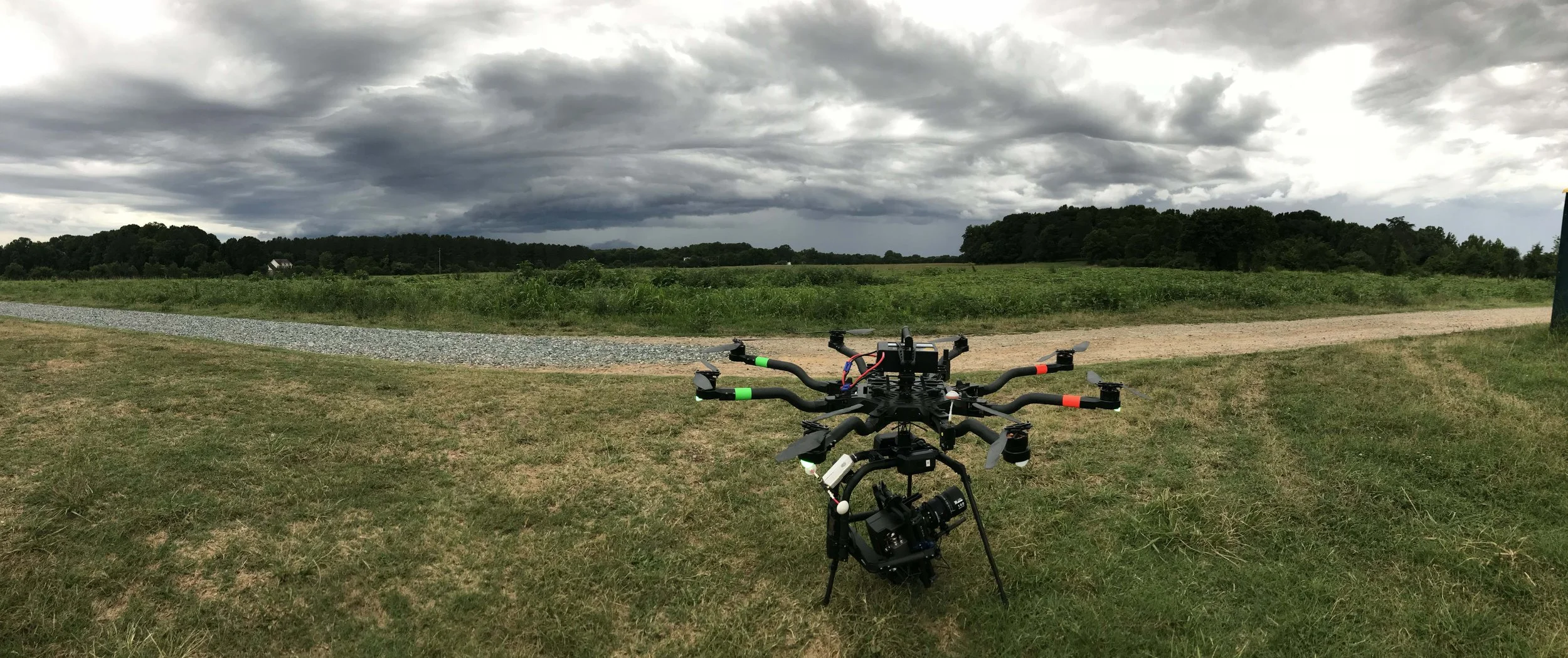 A large drone with multiple rotors is positioned on a grassy field overlooking a rural landscape with green fields, gravel and dirt paths, and a dense tree line. Dark clouds cover the sky, suggesting an approaching storm.