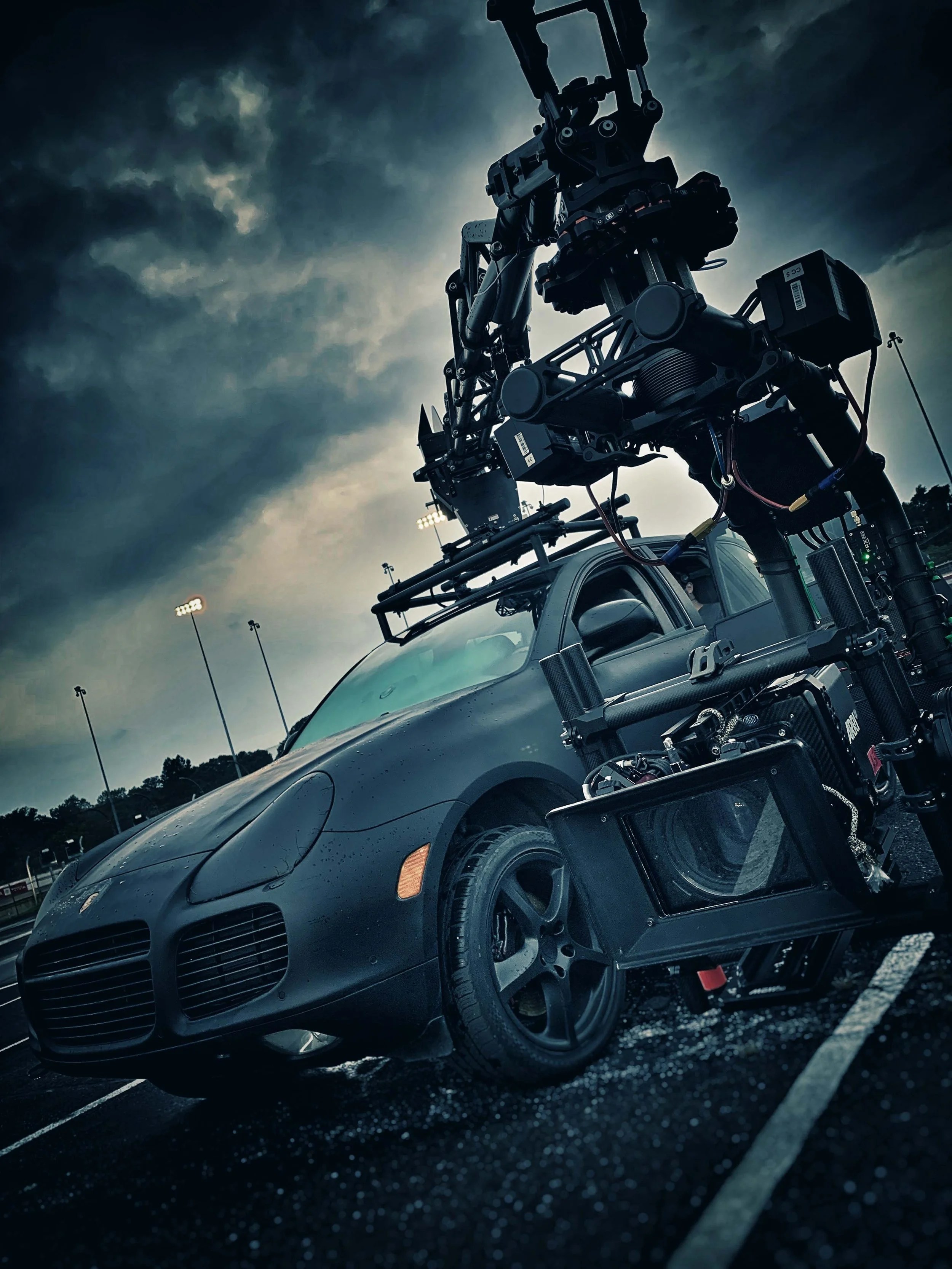 A film or television production camera mounted on a matte black sports car, set against a dark, overcast sky at a parking lot.