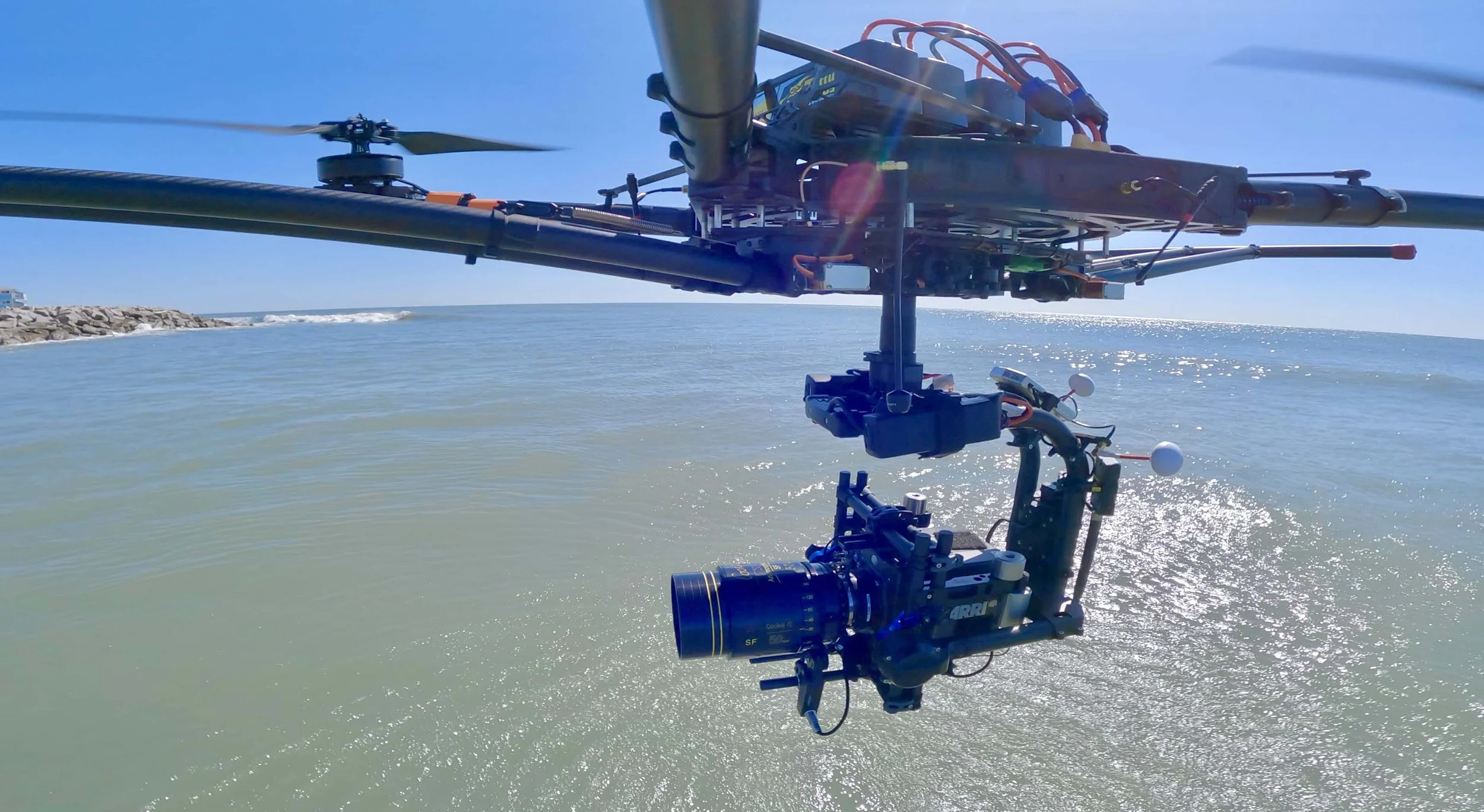 Close-up of a drone hovering above the ocean with a camera attached, with a rocky breakwater and blue sky in the background.