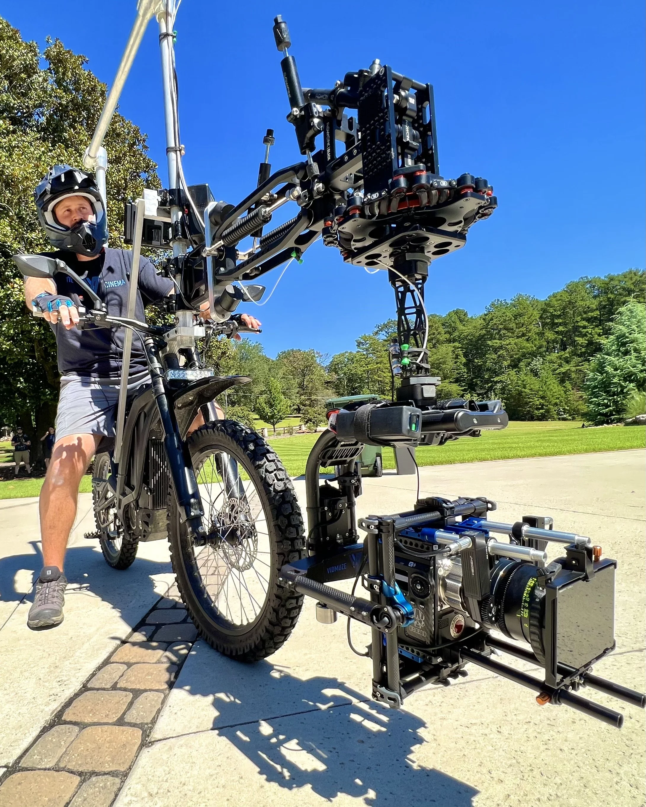 A man in a helmet and casual clothing rides an electric mountain bike equipped with a large professional camera mounted on a stabilization rig, set outdoors on a bright, sunny day.