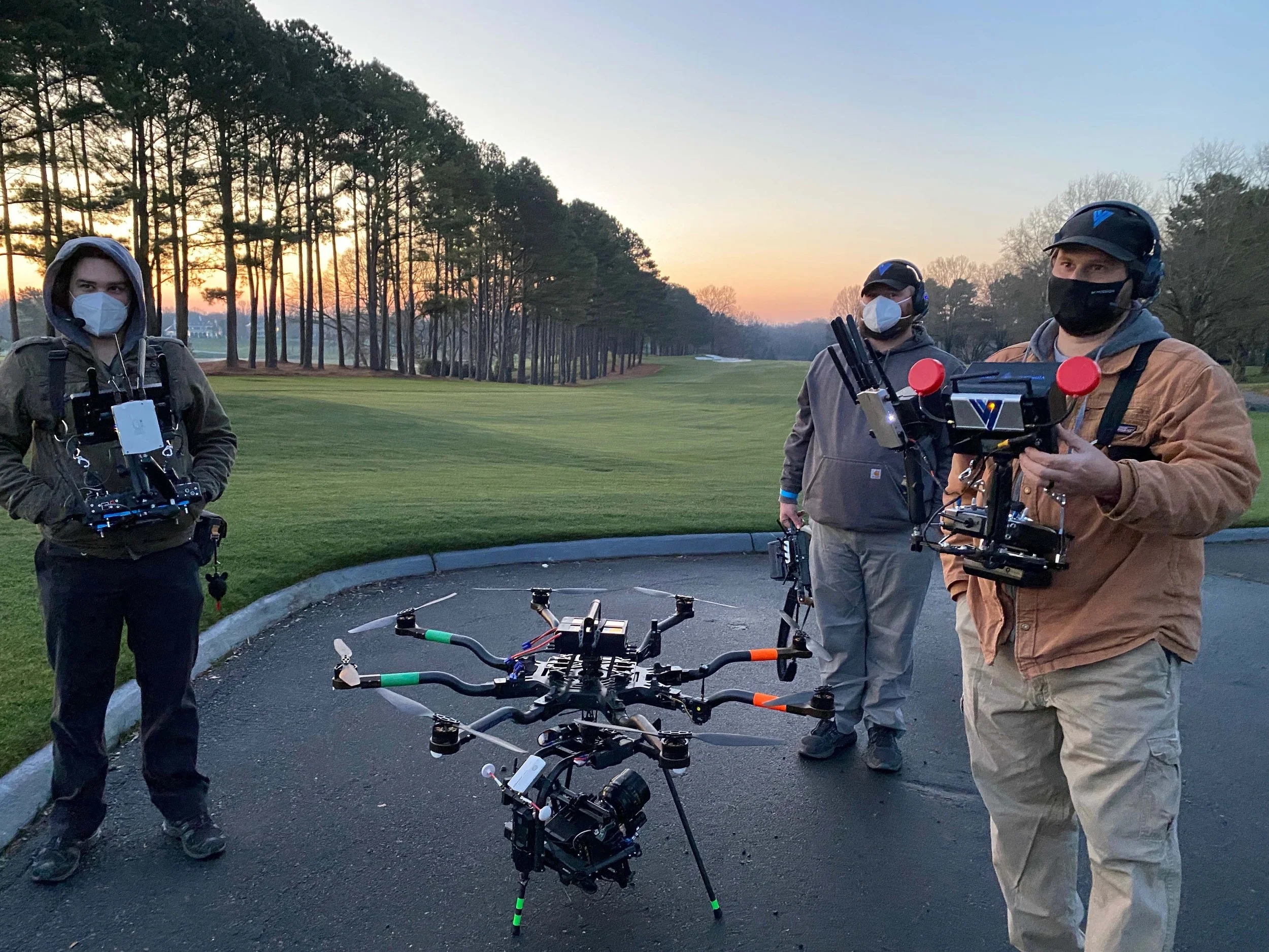 Three drone operators standing outdoors on a paved path at sunset, with a large drone on the ground and trees in the background.