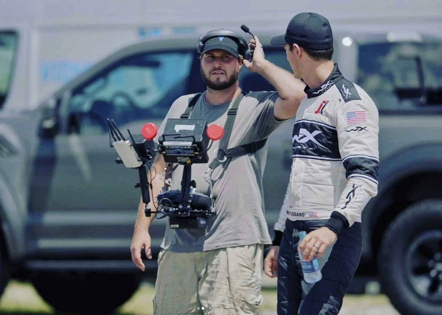Two men, one wearing a racing suit with an American flag patch, the other in casual clothing, are standing outdoors near a parked vehicle. They appear to be engaged in conversation, with one man adjusting a headset and the other holding a water bottl