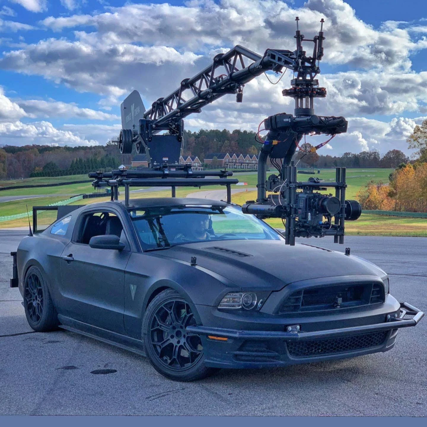 A matte black Ford Mustang with an advanced camera rig mounted on top, parked outdoors on a racetrack with a scenic background of fields, trees, and clouds.