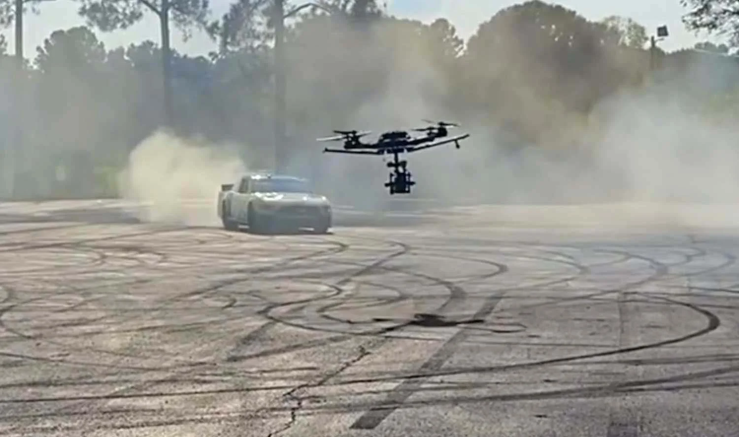 A drone flying near a police car on a cracked pavement with tire marks, surrounded by smoke or dust, possibly during a stunt or training exercise.