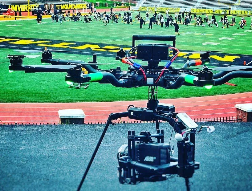 Drone placed on a tracked platform in front of a sports field with people and athletes in the background.
