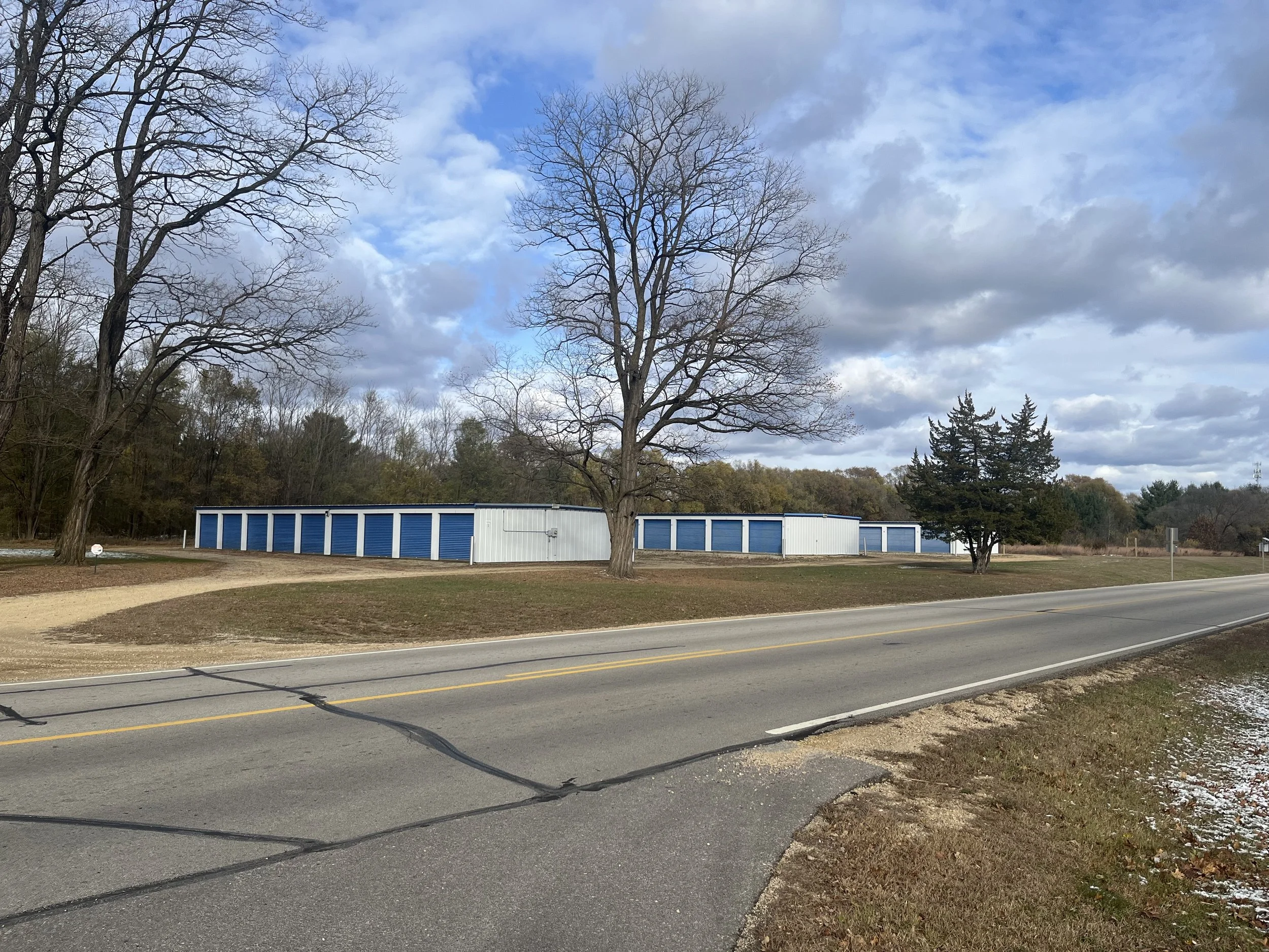 A rural area with a paved two-lane road, leafless trees, and a white building with blue doors in the background. The sky is partly cloudy.