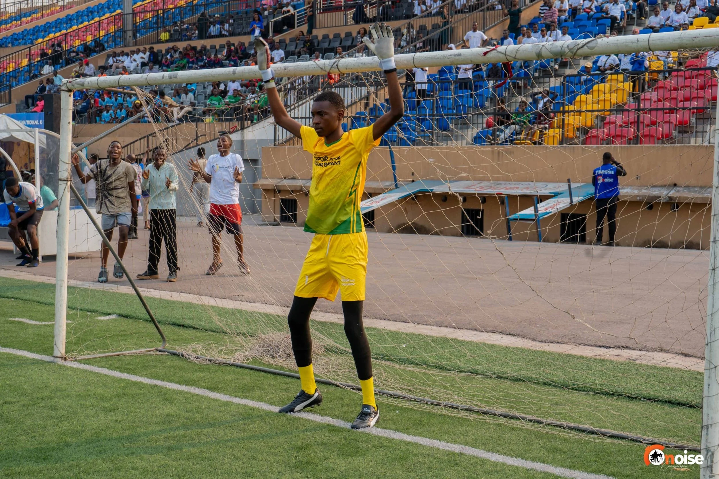 A soccer goalkeeper wearing a yellow and green uniform standing in front of the goal net with arms raised. Several spectators are seated in the stands behind. The scene is at a soccer stadium.
