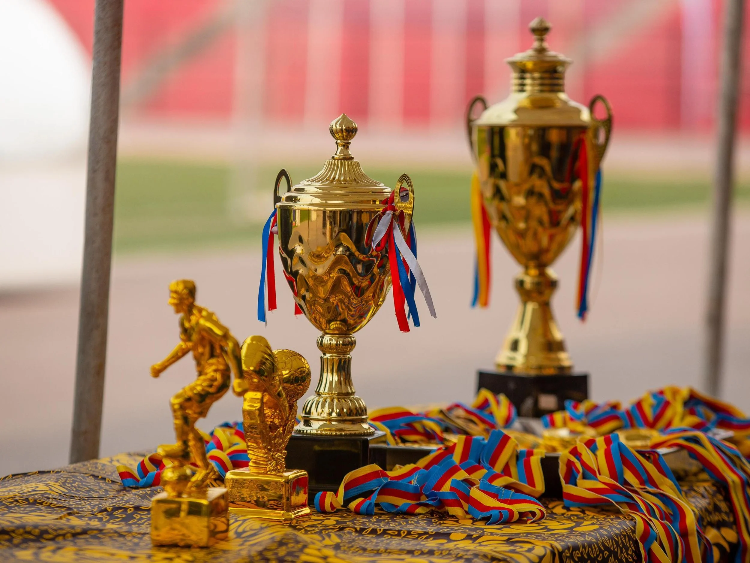 Gold trophies and medals displayed on a table at a sports event, with red, yellow, blue, and white ribbons attached to the trophies and medals.