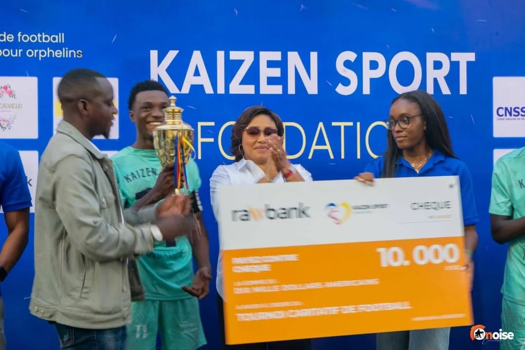 Group of young men receiving a trophy and large check for 10,000 from woman at an award ceremony. Blue background with 'KAIZEN SPORT FOUNDATION' text. One man in teal shirt is smiling, another in beige jacket is clapping.