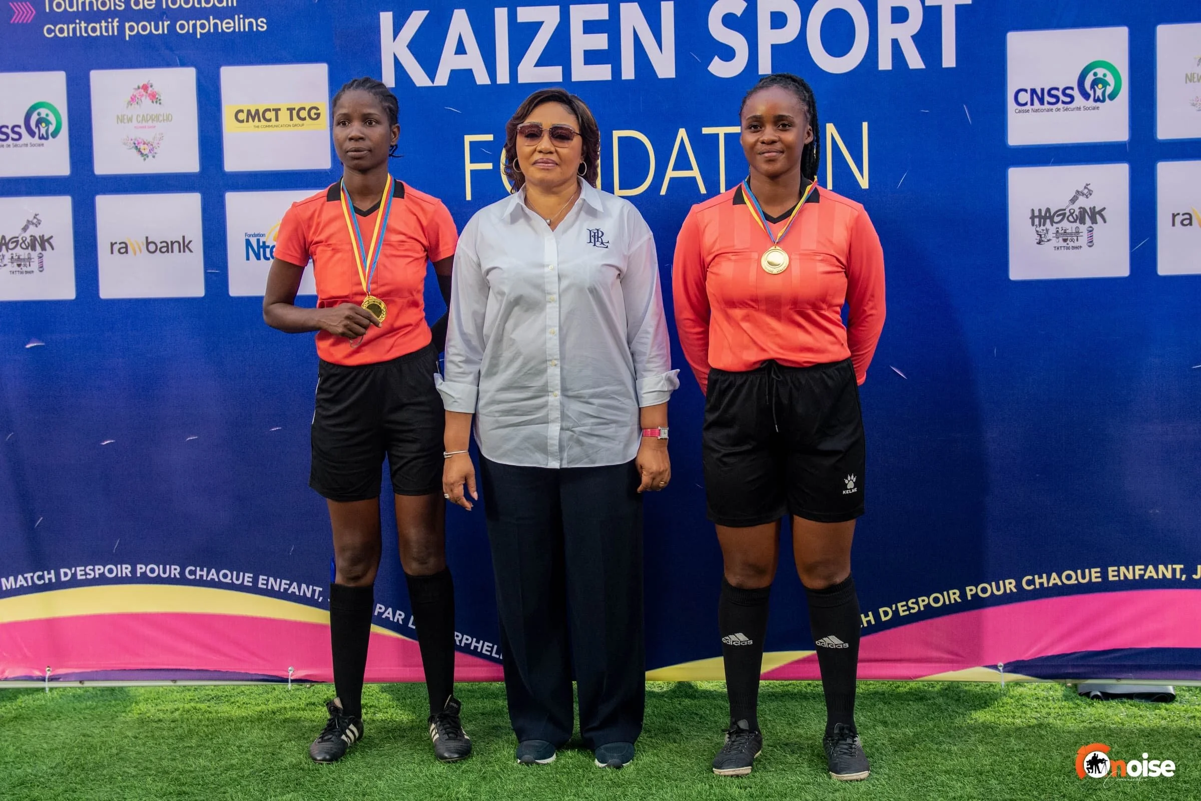 Two female athletes dressed in orange shirts and black shorts wearing medals, standing beside a woman in white shirt in front of a blue backdrop with various sponsor logos and event information at a sports award ceremony.