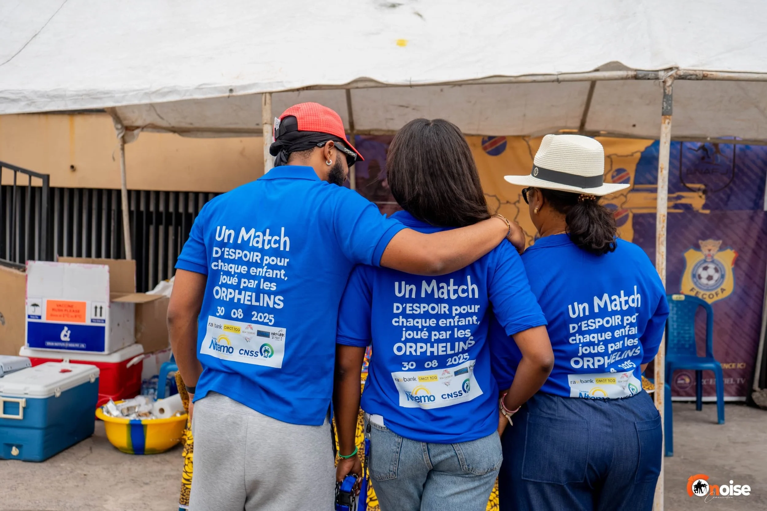 Three people wearing blue T-shirts with white text, standing together with their backs to the camera at an outdoor shelter.