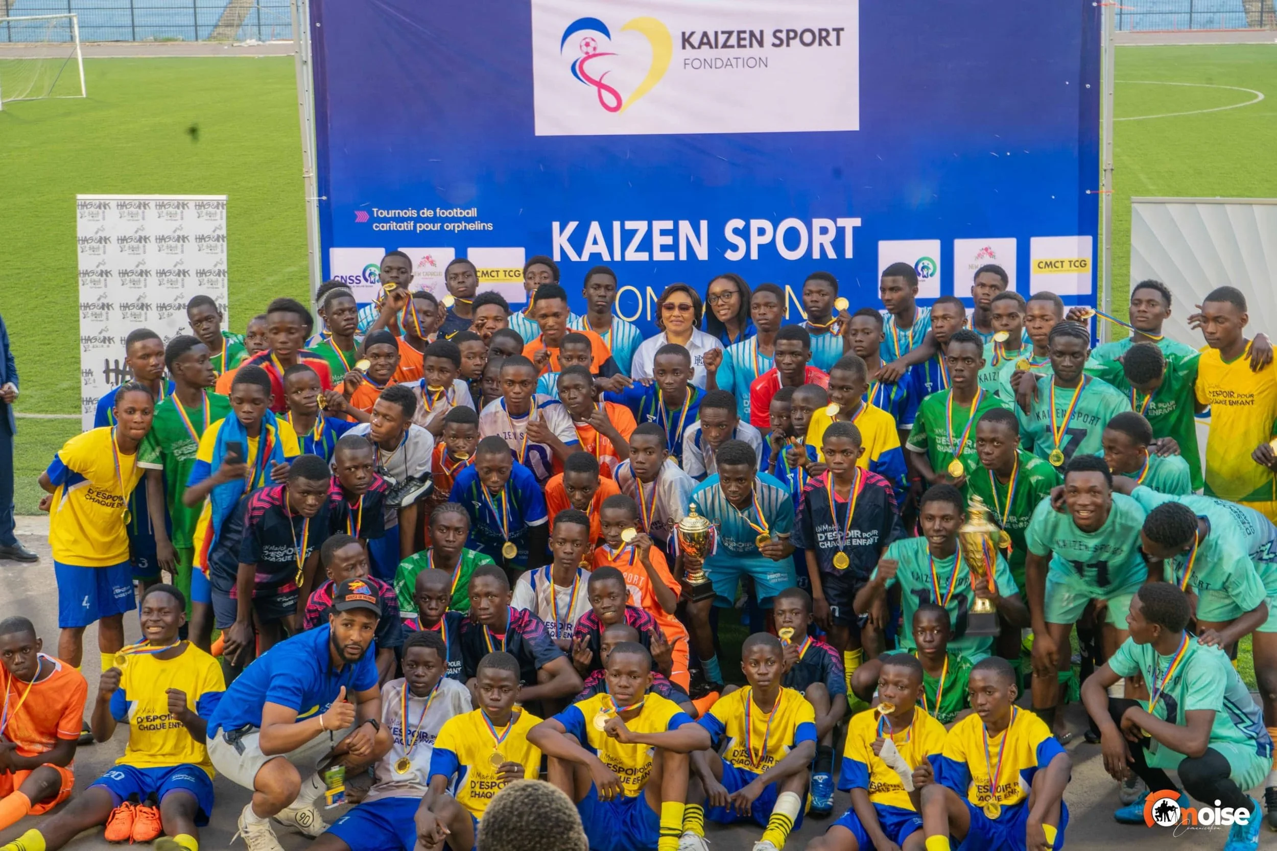 Group of young soccer players and coaches posing with trophies and medals on a soccer field, celebrating their victory at a sport event. Behind them is a large blue banner with the text 'KAIZEN SPORT' and logos, including a heart-shaped logo with a s