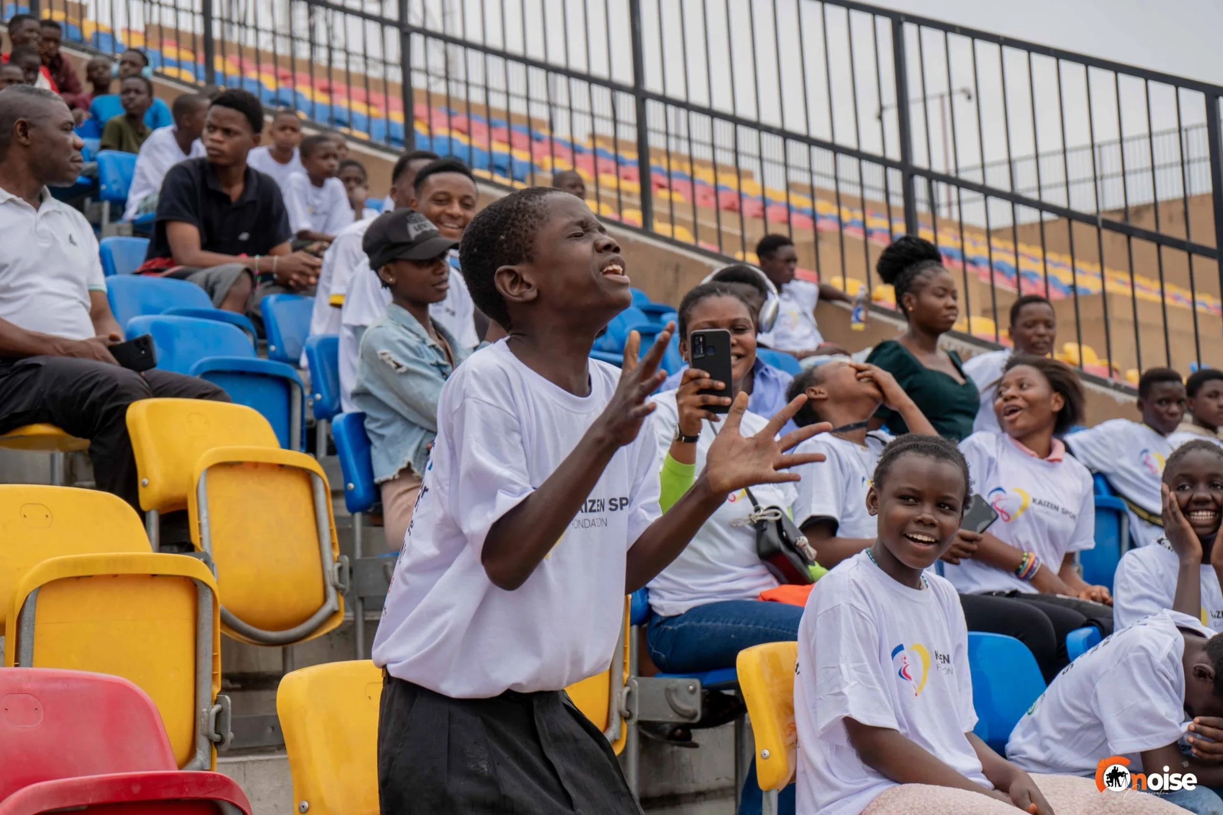 Young girl in white t-shirt appears to be singing or shouting with hands raised, surrounded by other children and adults sitting in colorful stadium seats, some smiling, at a sports event or gathering in an outdoor stadium.
