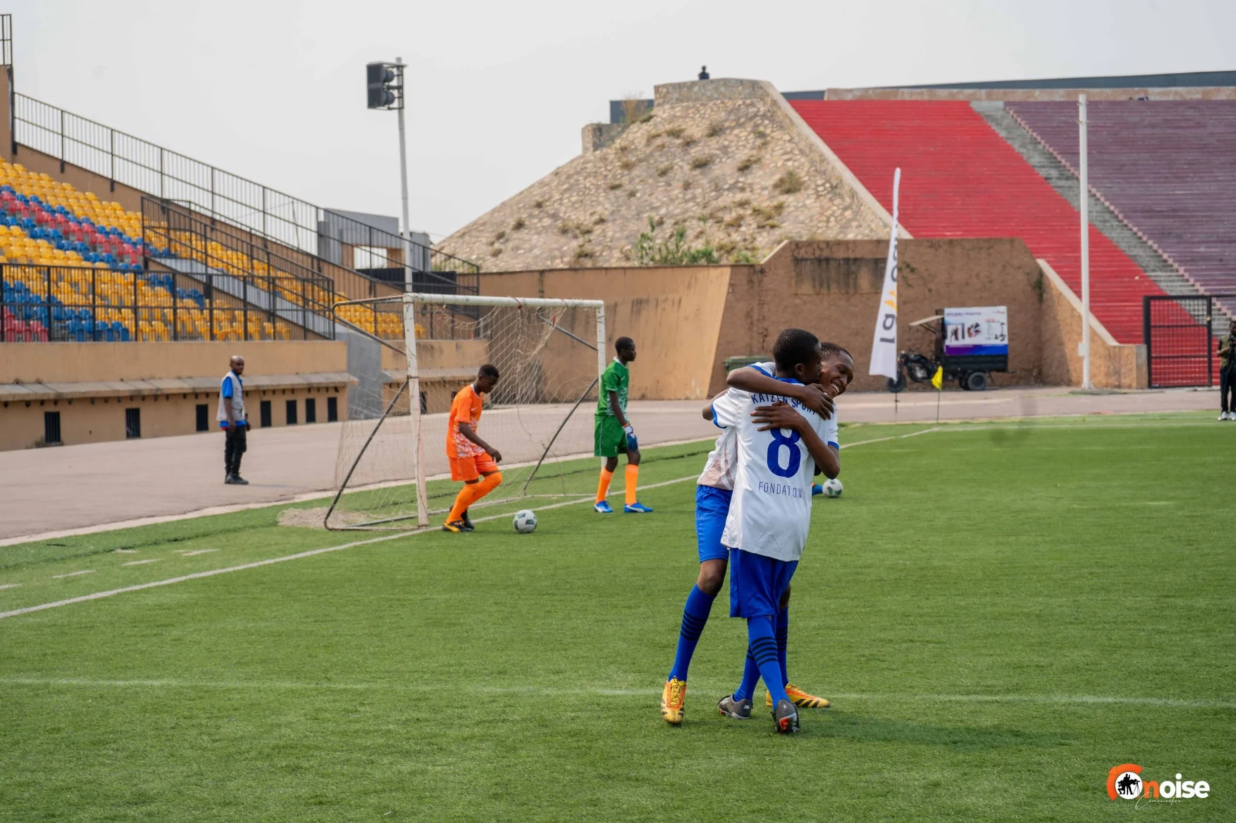 Two young soccer players in blue uniforms hugging each other on the field, celebrating. In the background, a soccer goal with two children in orange and green goalkeeper uniforms near the goal, and a person in a blue jacket walking along the track. E