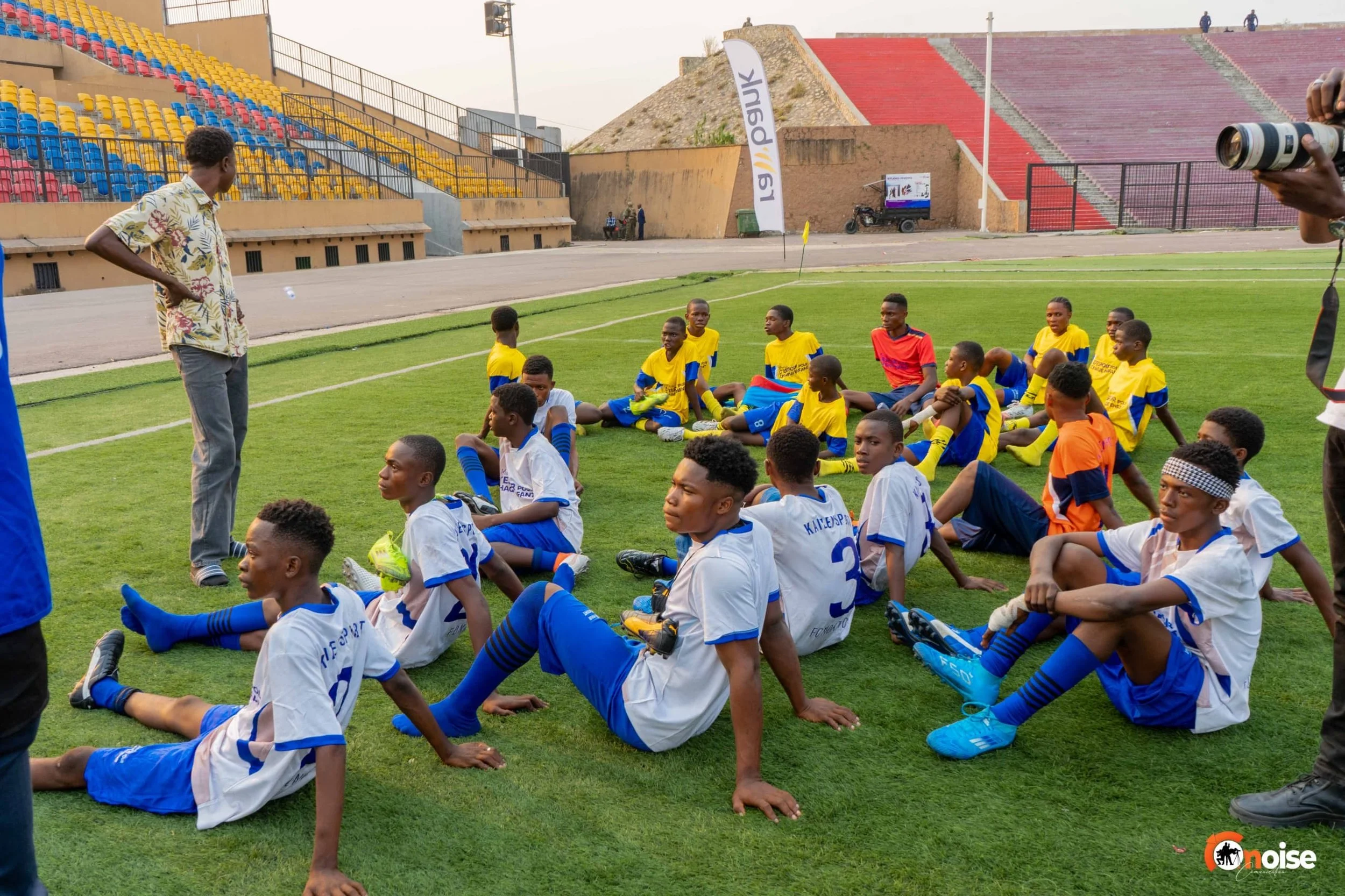 Young soccer players sitting on the field listening to their coach during a training session at a stadium with red and yellow seats.