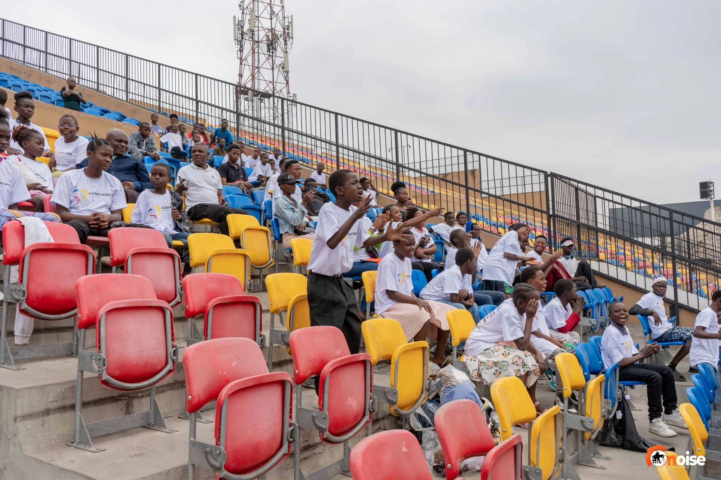 Children and adults seated in colorful stadium seats, some standing and gesturing, during an outdoor event on a cloudy day.