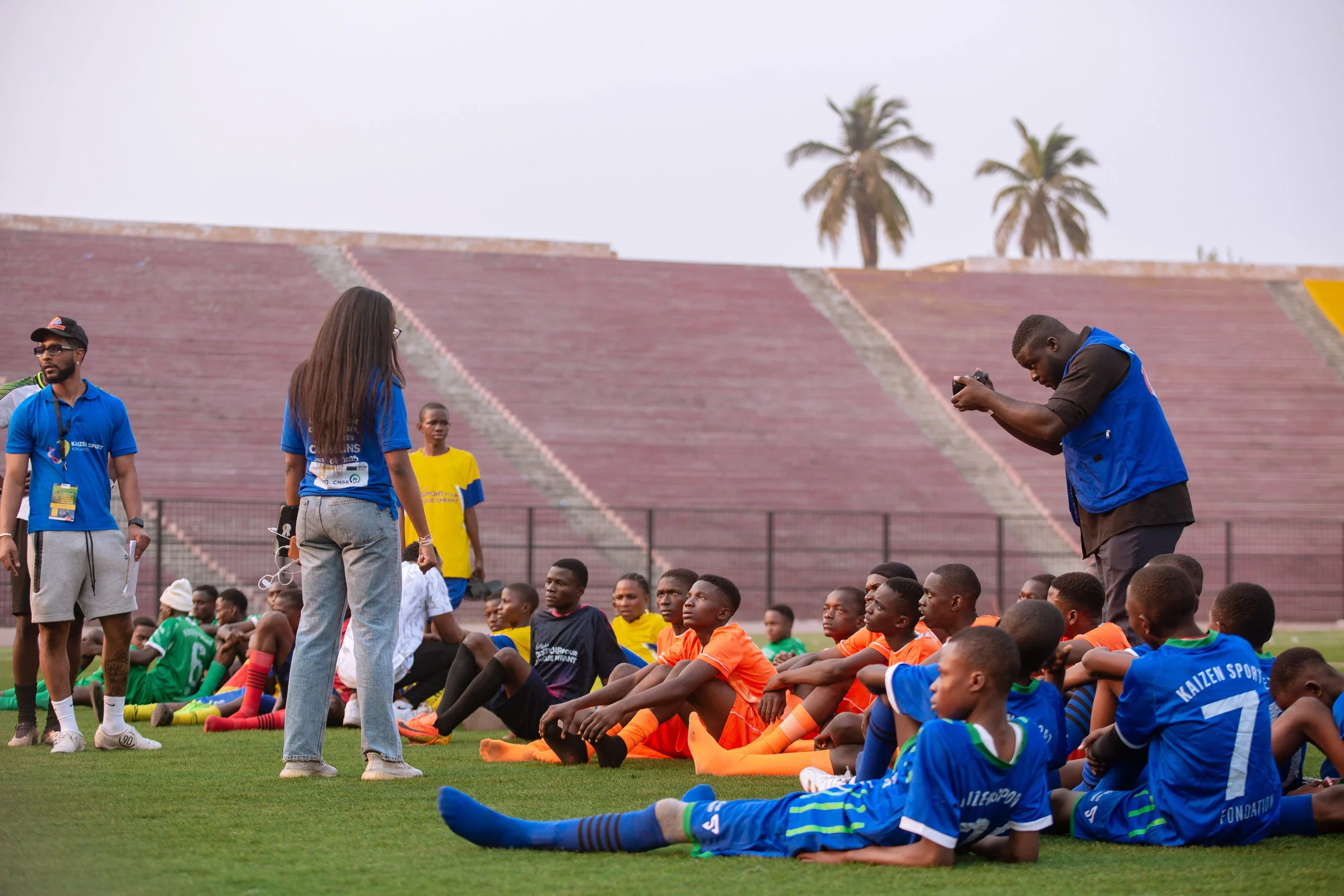 Youth soccer players seated on the grass during a training session or game, with coaches and adults standing nearby, in a stadium with palm trees in the background.
