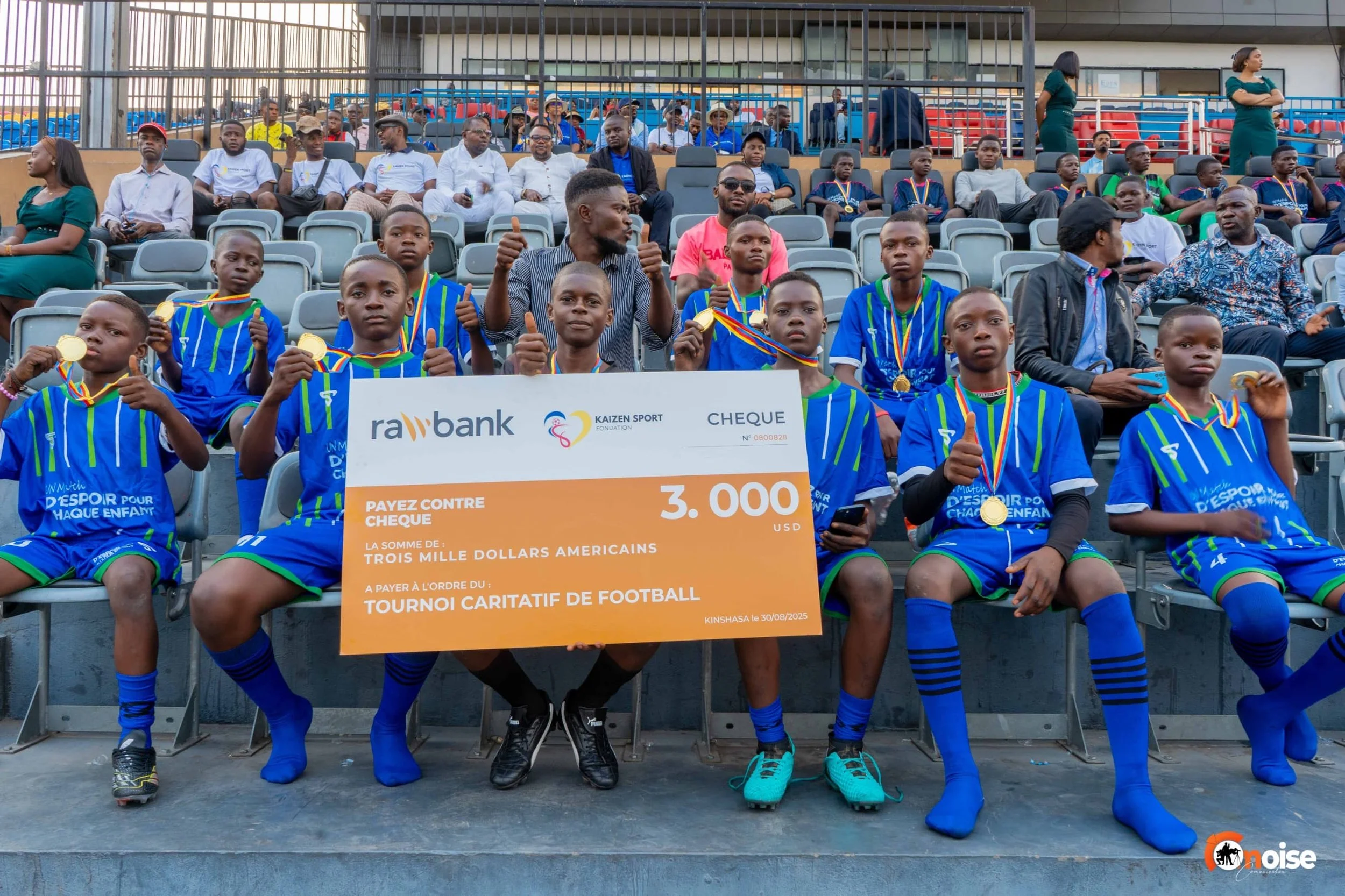 Children in blue football uniforms holding a large check for $3,000 and medals, sitting in stadium seats after a charity football tournament in Kinshasa, August 30, 2025.