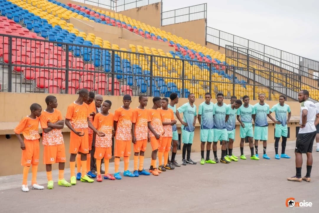 A group of young soccer players in orange and teal uniforms standing in a line on a soccer field while listening to a coach.