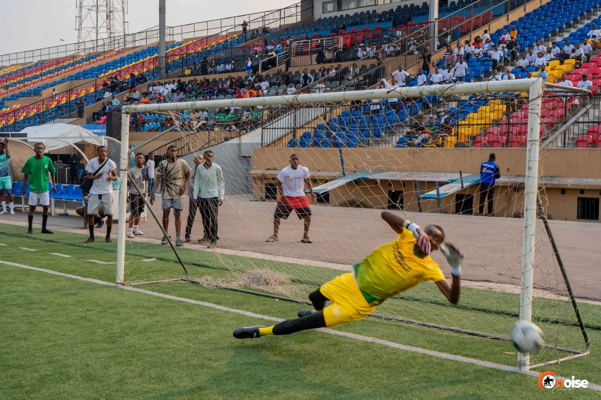 A soccer goal with a goalkeeper in yellow uniform diving to the right to save a ball, while players and spectators watch in a stadium with colorful seats.