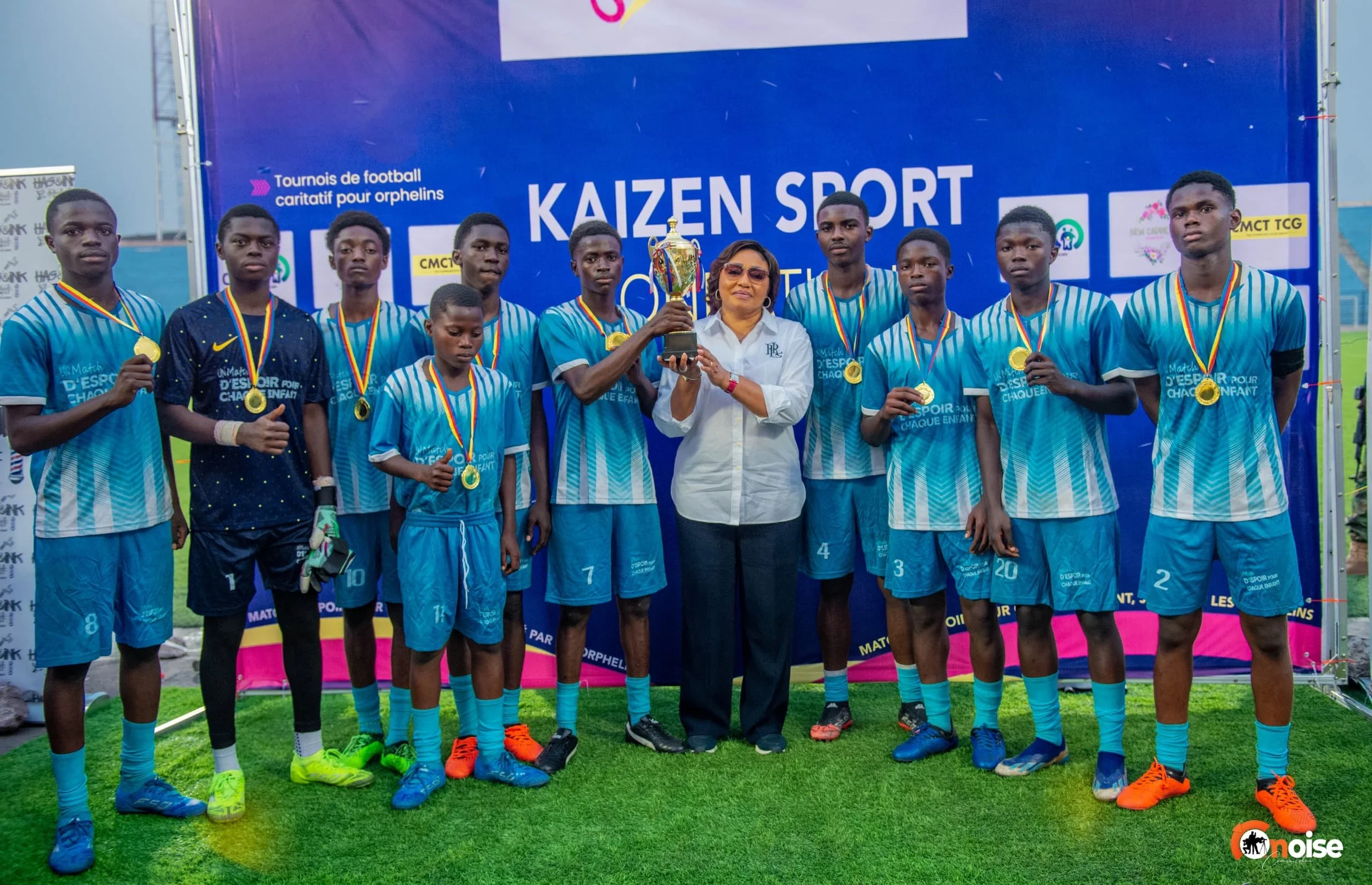 A youth soccer team poses with medals and a trophy after a match or tournament victory, with a woman in sunglasses at the center holding the trophy.