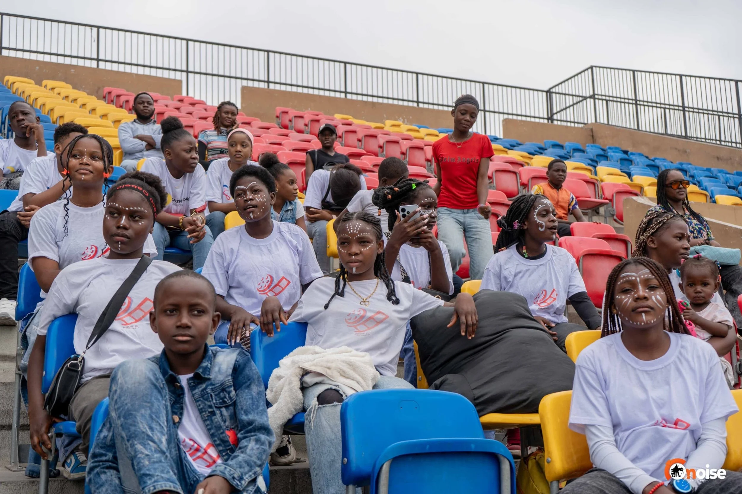 Group of diverse children and adults sitting in stadium bleachers, some with face paint and wearing matching t-shirts, with a woman standing nearby using a smartphone.