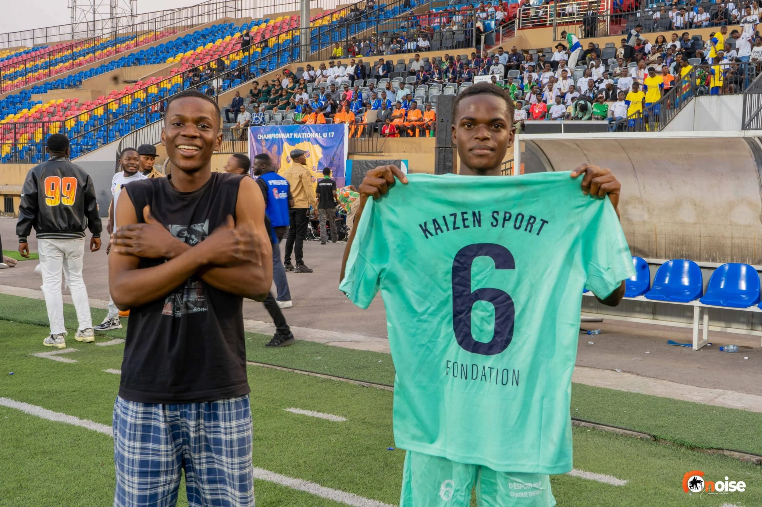 Two young men on a football field, one holding a light green jersey with the text 'KAIZEN SPORT 6 FONDATION' and the other with arms crossed, smiling, with a crowded stadium and spectators in the background.