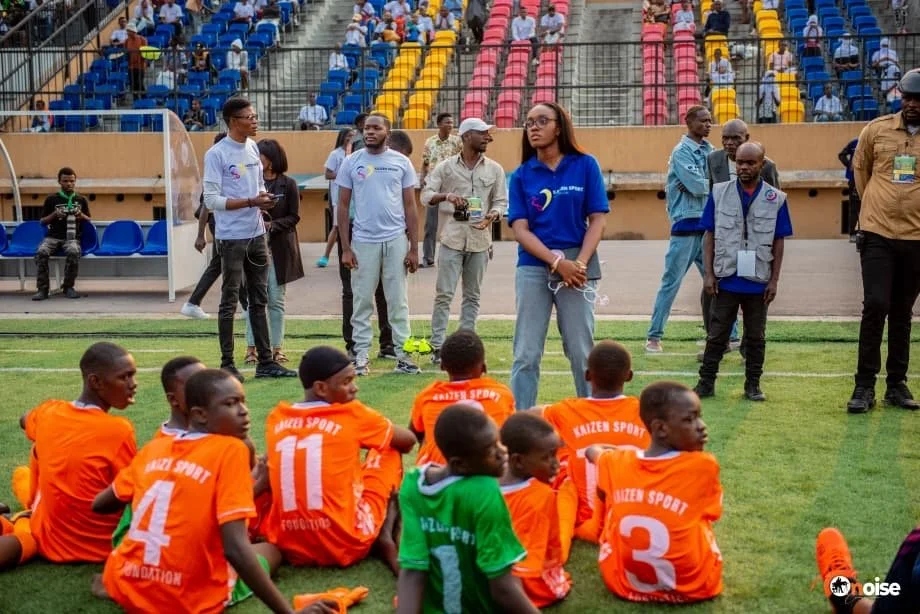 Young soccer players in orange jerseys sitting on the grass at a stadium, with coaches and spectators in the background.