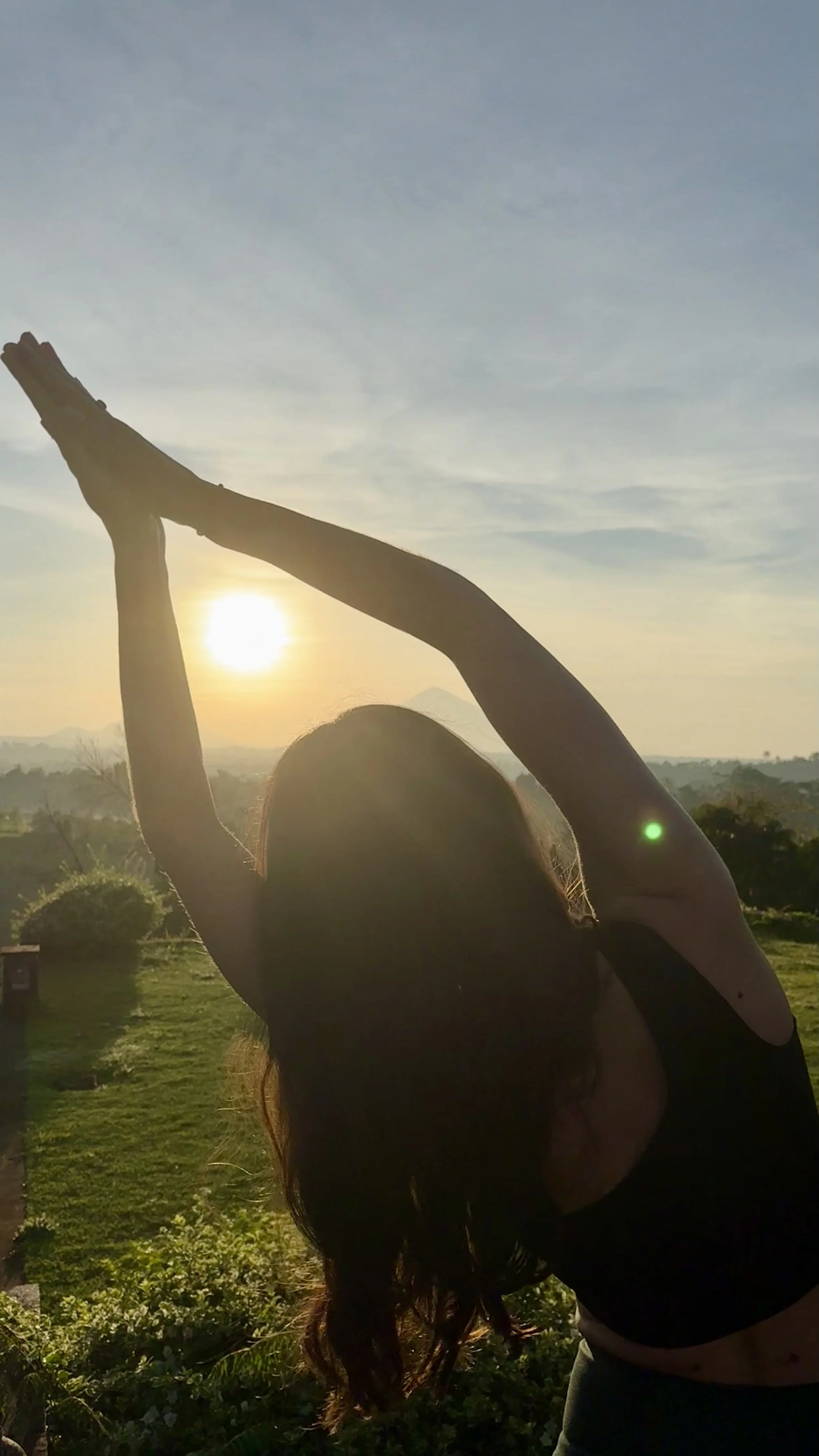 Frau praktiziert Yoga im Freien bei Sonnenaufgang mit Blick auf Himmel und grüne Landschaft.