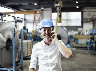 A man in a white shirt and blue safety helmet smiling and talking on a mobile phone inside an industrial factory setting.